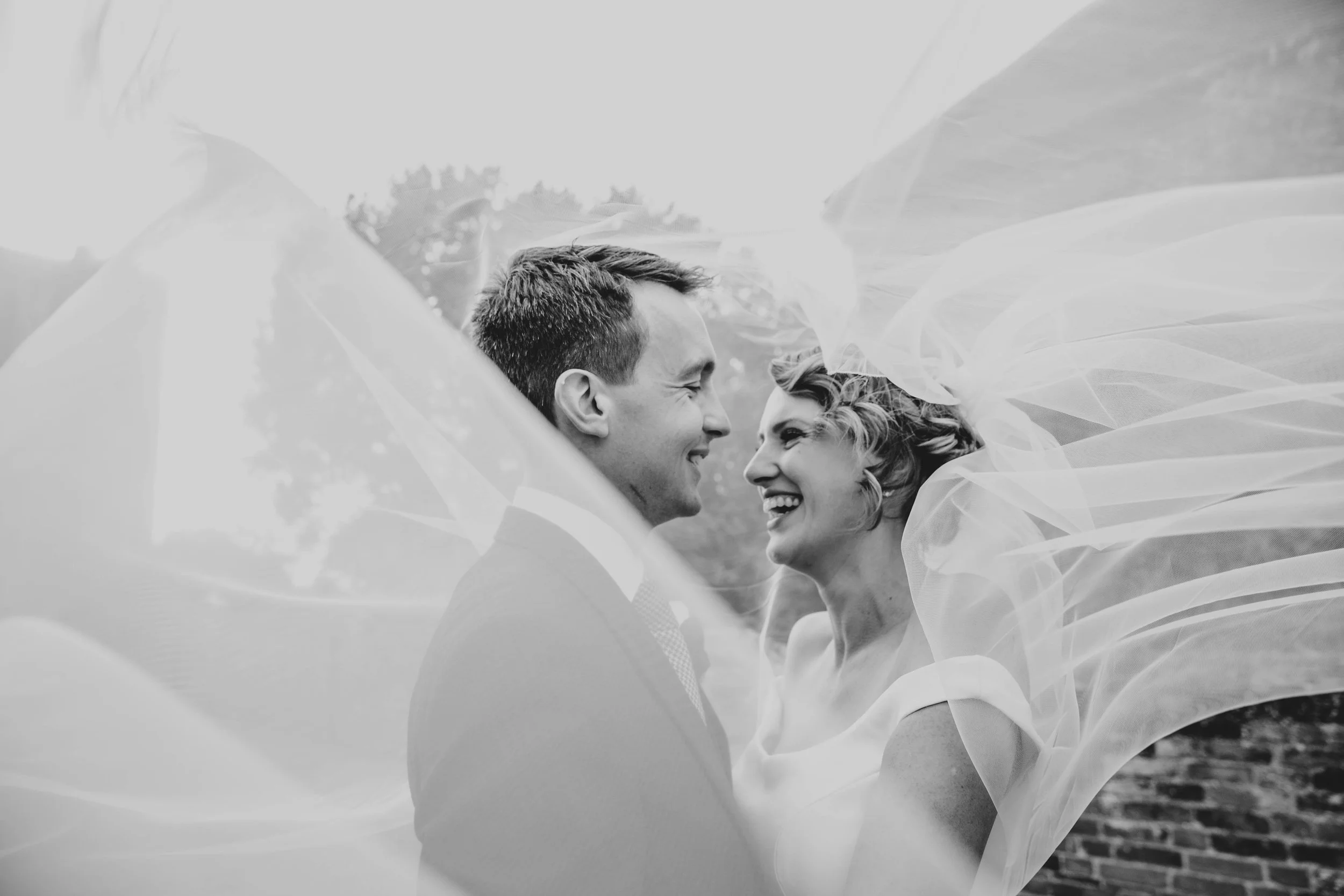 Bride &amp; Groom laugh under windswept veil in Hedingham Castle Wedding Essex Documentary Wedding Photographer