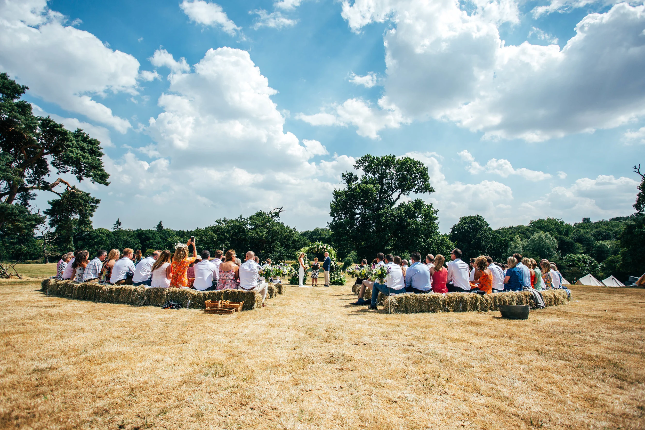 Guests sit on hay bales under fluffy clouds for diy back garden wedding Essex Documentary Wedding Photographer