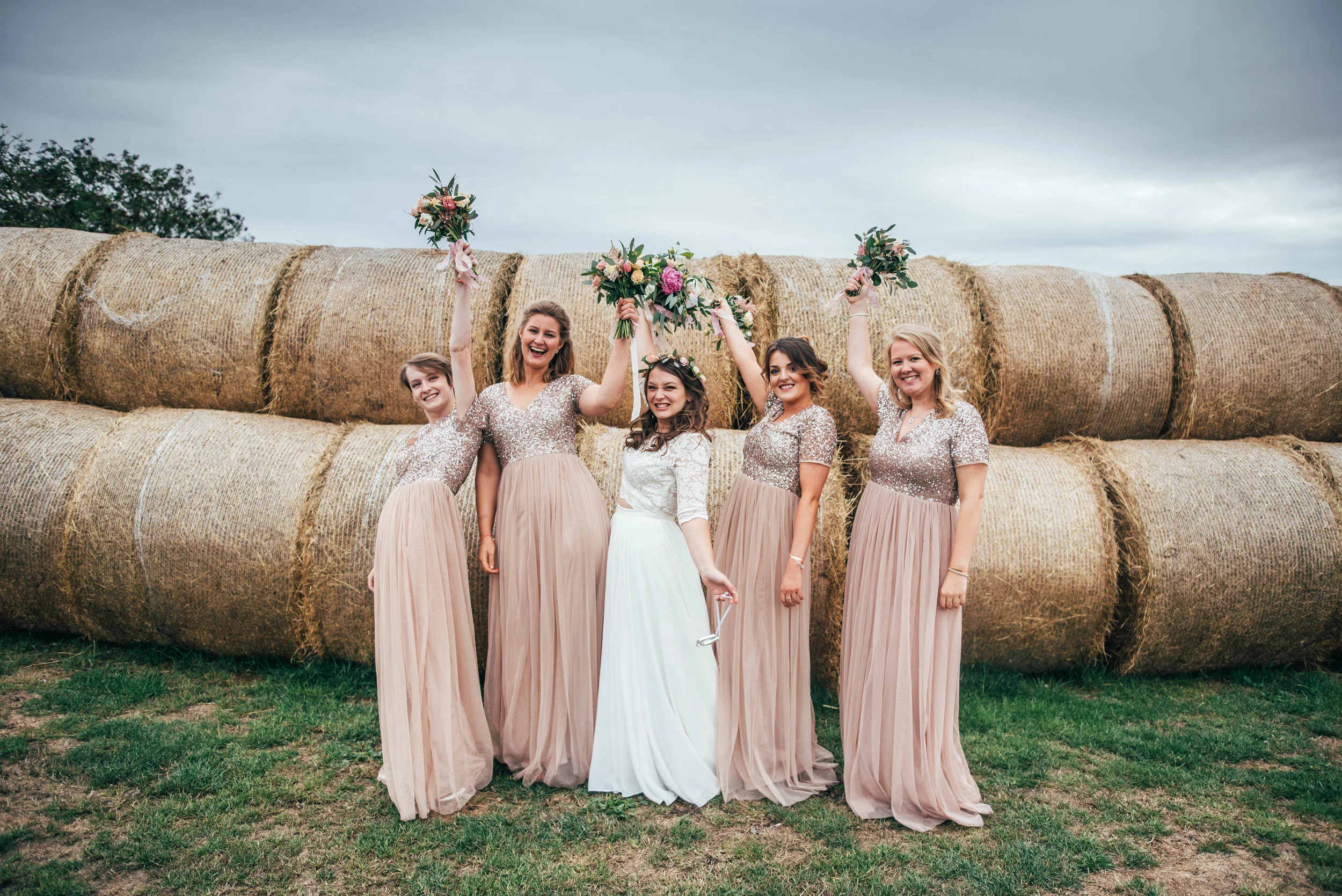 Bride and Bridesmaids lift bouquets by hay bales at Bedinghams Farm, Ely.  Essex Documentary Wedding Photography