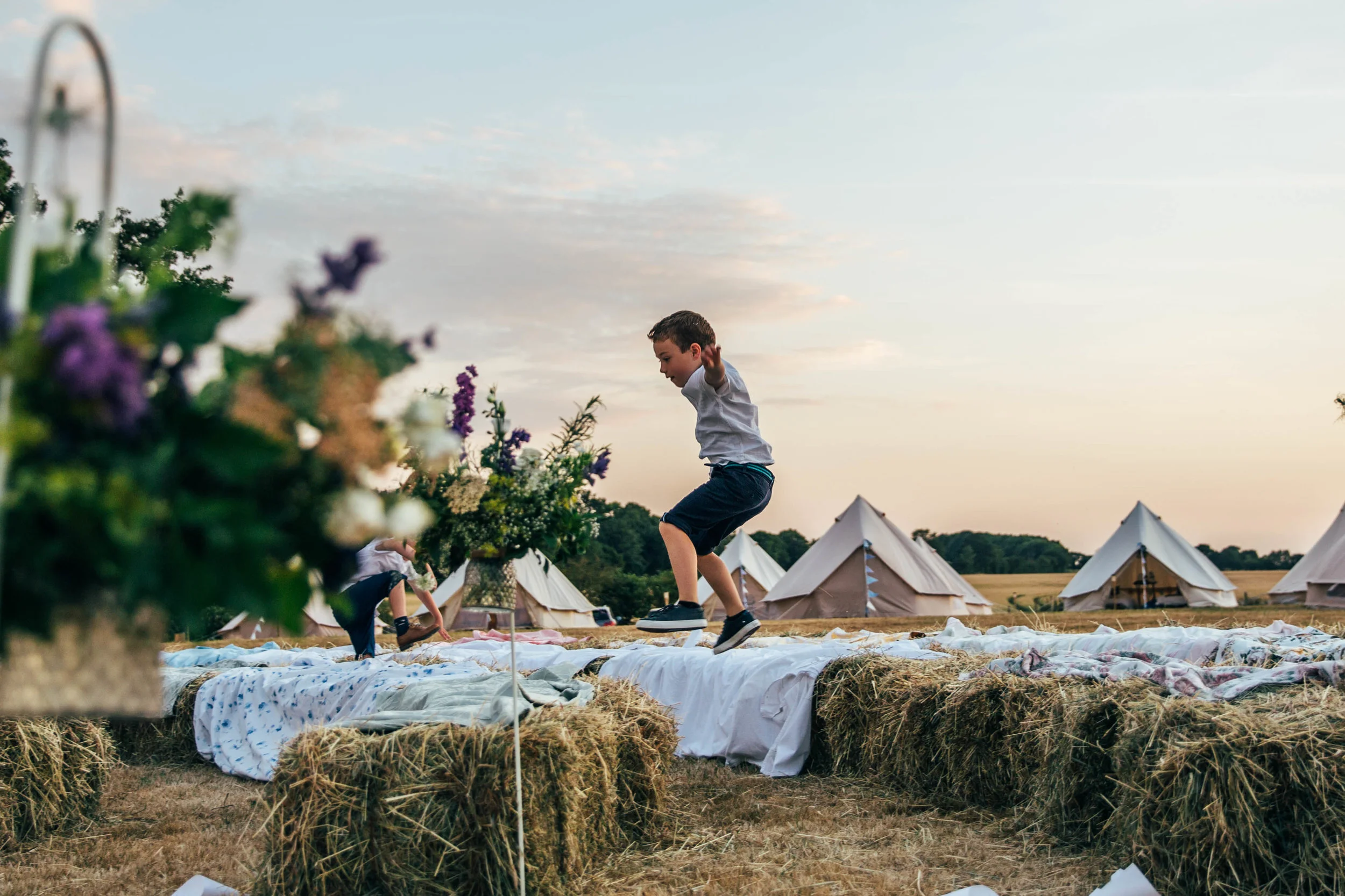 Little boy jumps over Hay Bale seats at sunset outdoor DIY wedding Essex Documentary Wedding Photographer