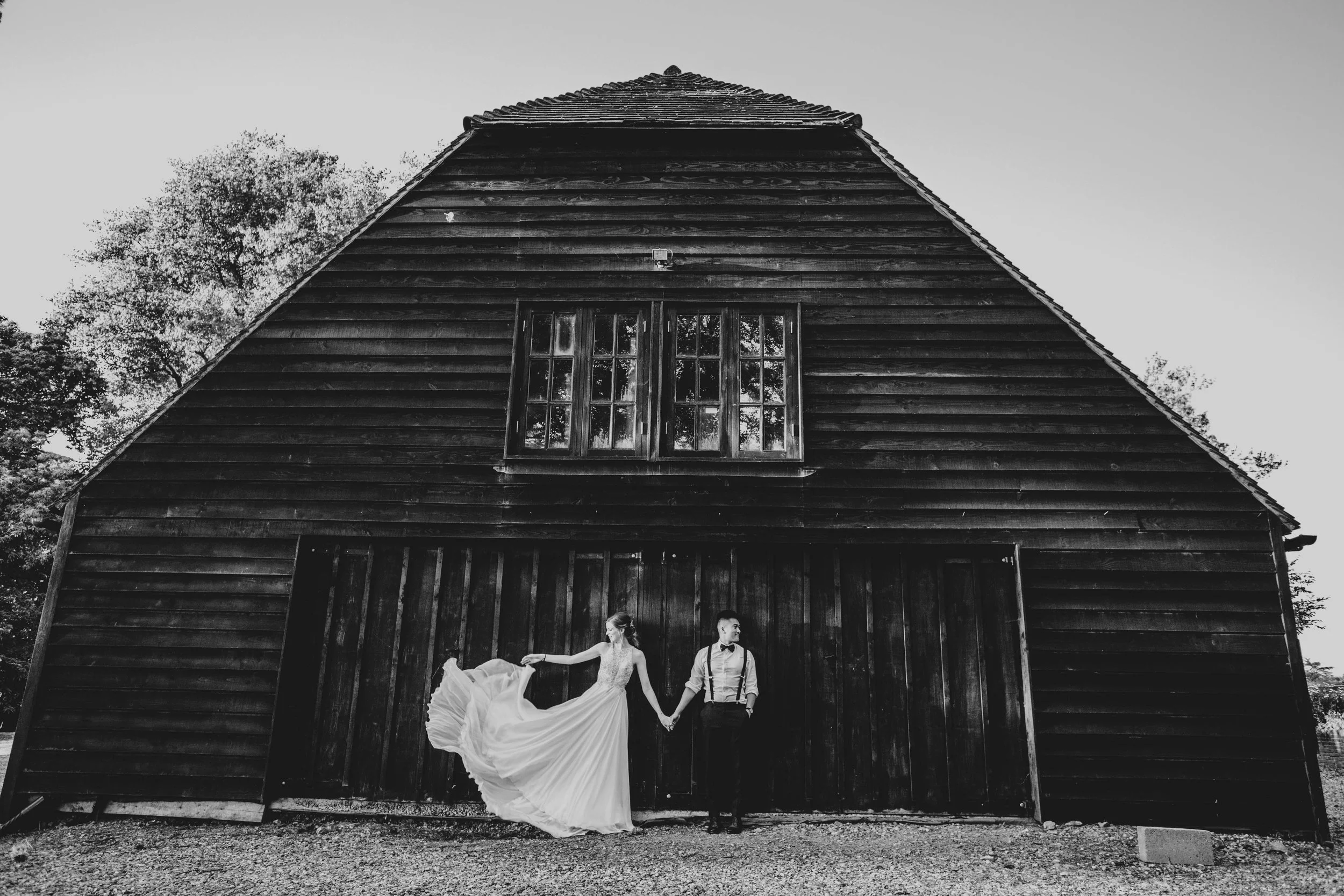 Bride with swish dress and Groom stand outside black barn at The Long Barn, Essex Documentary Wedding Photographer