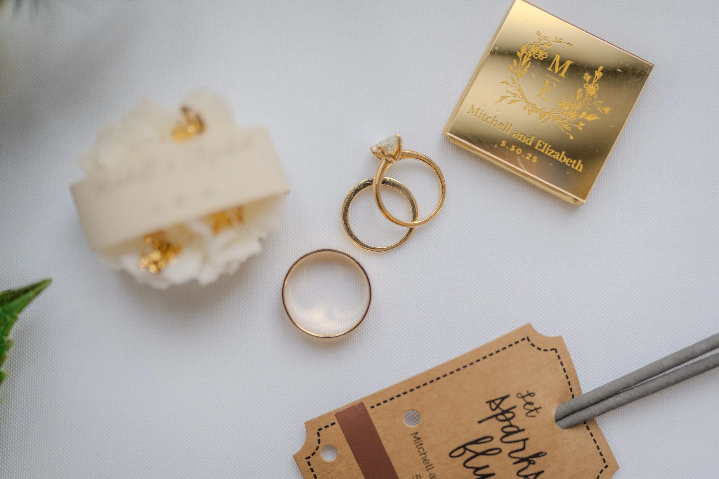 Wedding rings, wedding band, custom jewelry box, wedding invitation, and a small decorative flower on a white surface.
