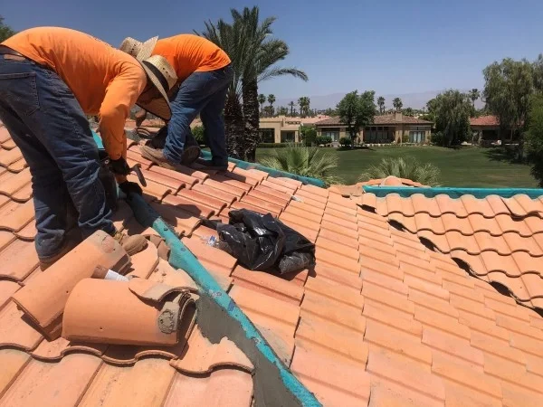 Roofing contractors replacing clay roof tiles on a residential home under clear sunny skies.