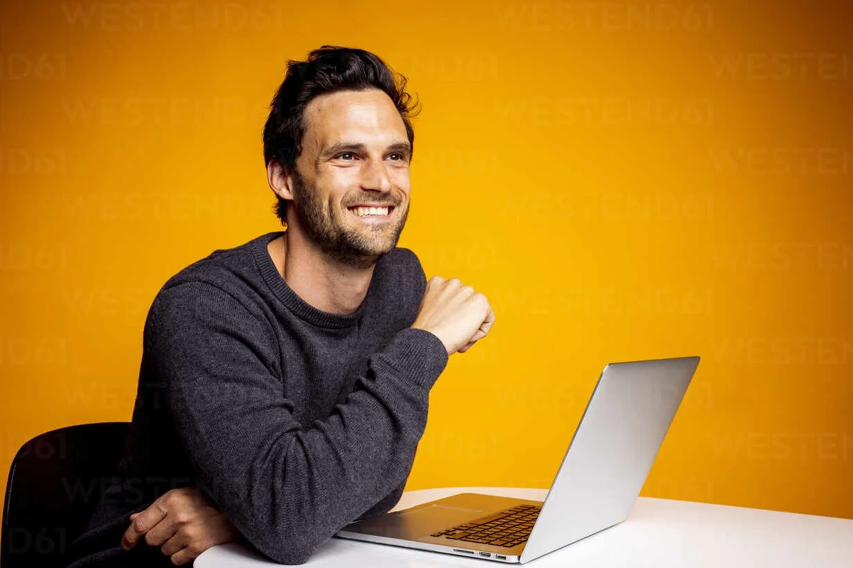 thoughtful-happy-man-looking-away-while-sitting-with-laptop-at-table-against-yellow-background-DAWF01620.jpg