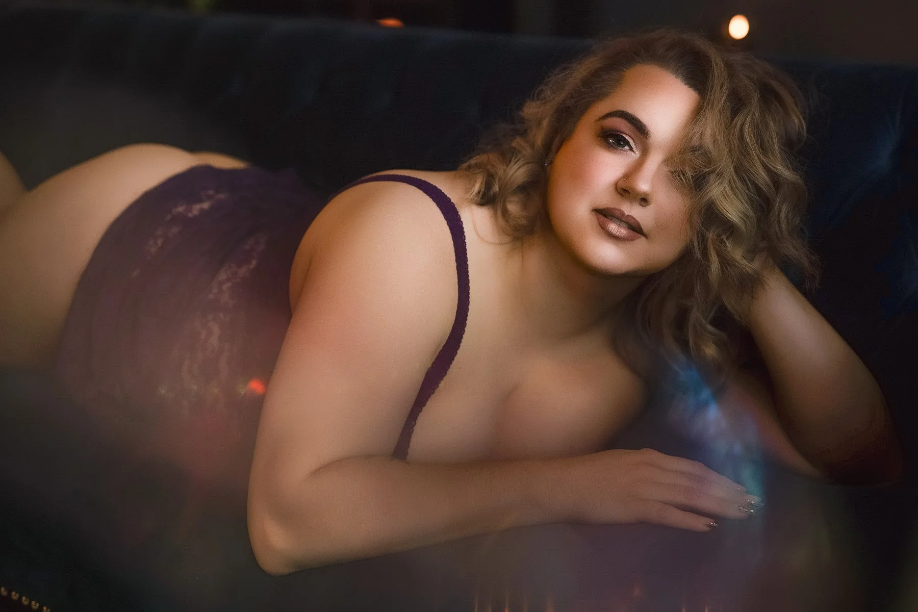 A woman in a dark lace bodysuit lies on her side on a dark couch, looking at the camera with a relaxed, confident expression.