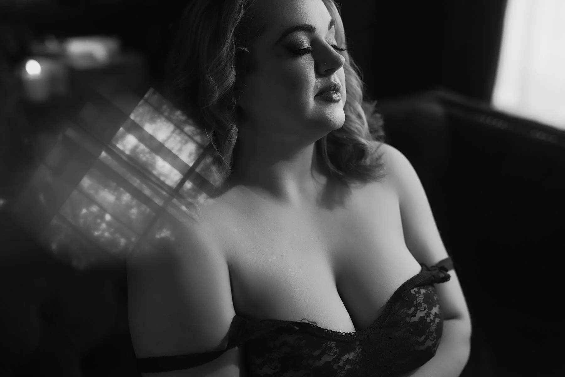 A woman in lace lingerie sits with eyes closed in soft, moody light; the image is in black and white.