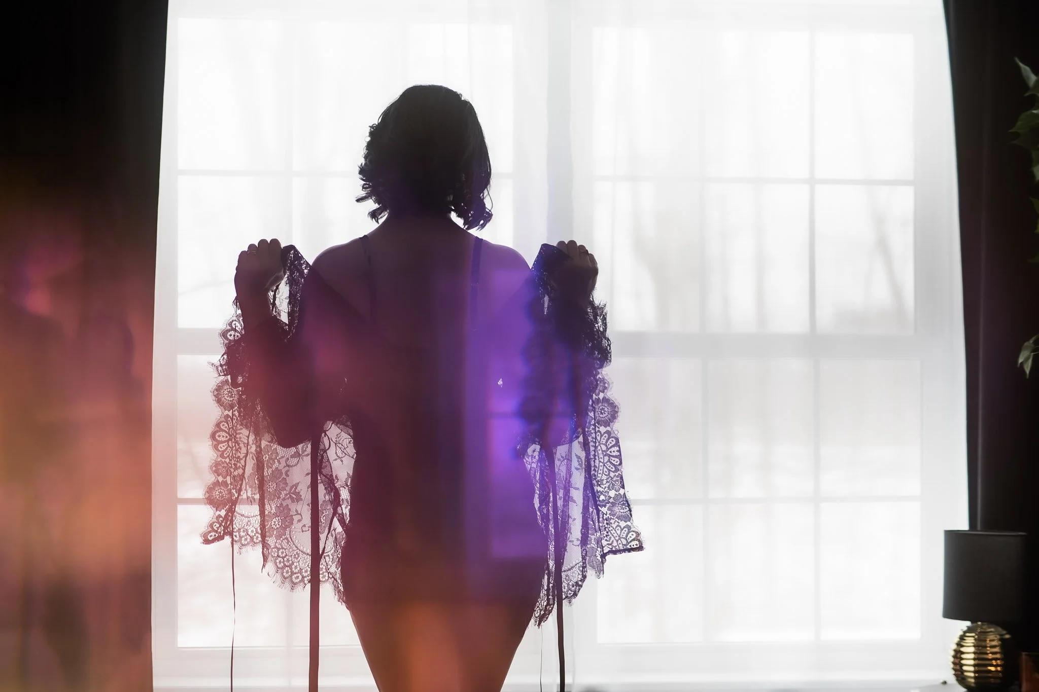 A person in lace lingerie stands facing a bright window, silhouetted by natural light, holding a sheer robe.