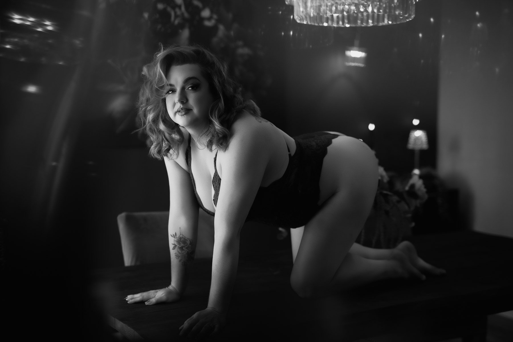 A woman in lingerie poses confidently on a table in a dimly lit, elegant room. Black and white photo