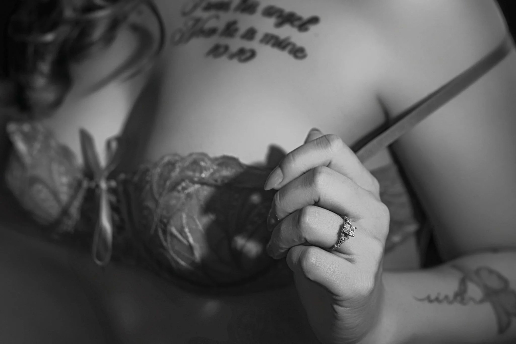A woman in lace lingerie holds a strap, displaying a diamond ring and tattoos across her chest and arm.