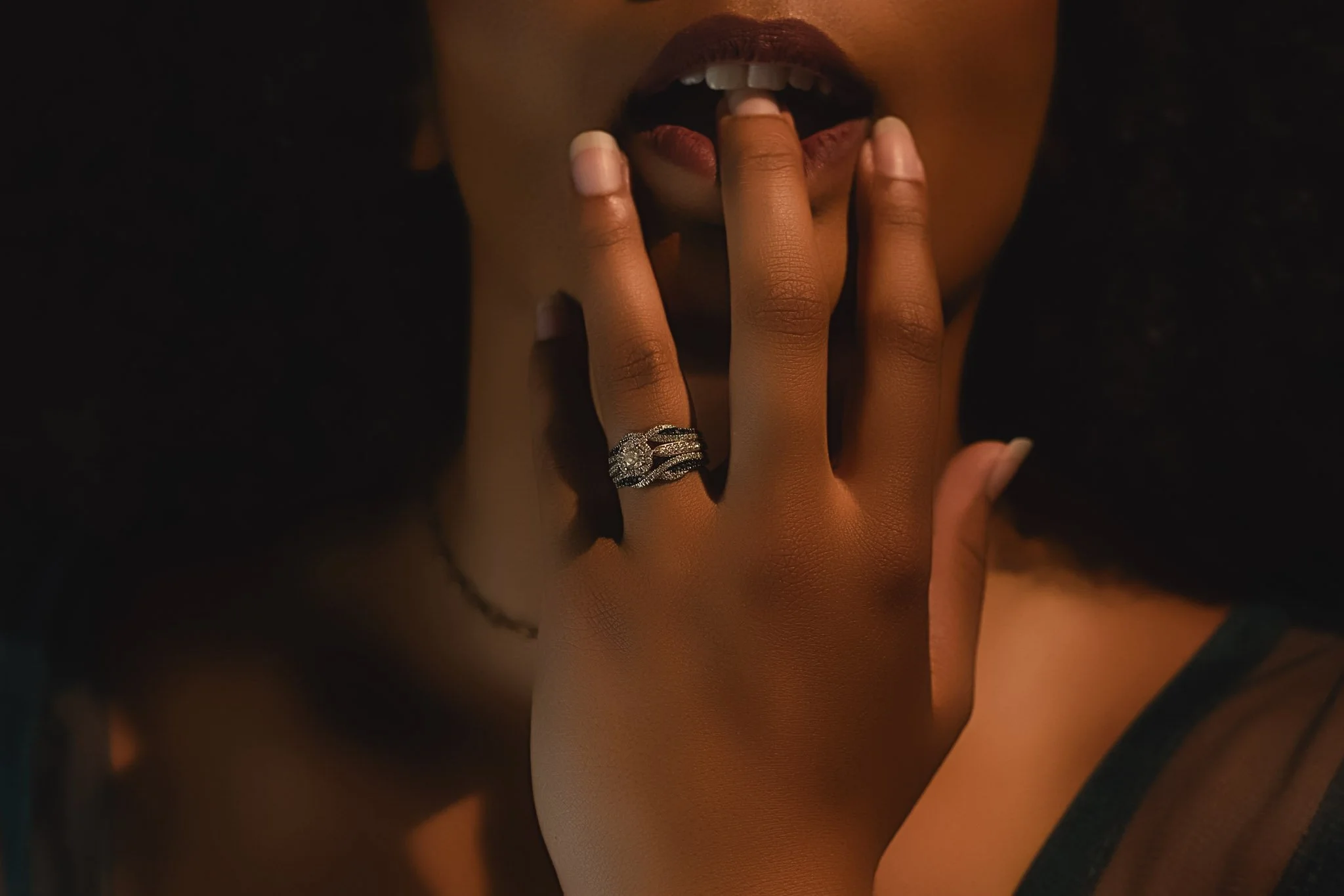 A person touches their lips with fingers, wearing a wedding ring on their finger in dim, moody lighting.