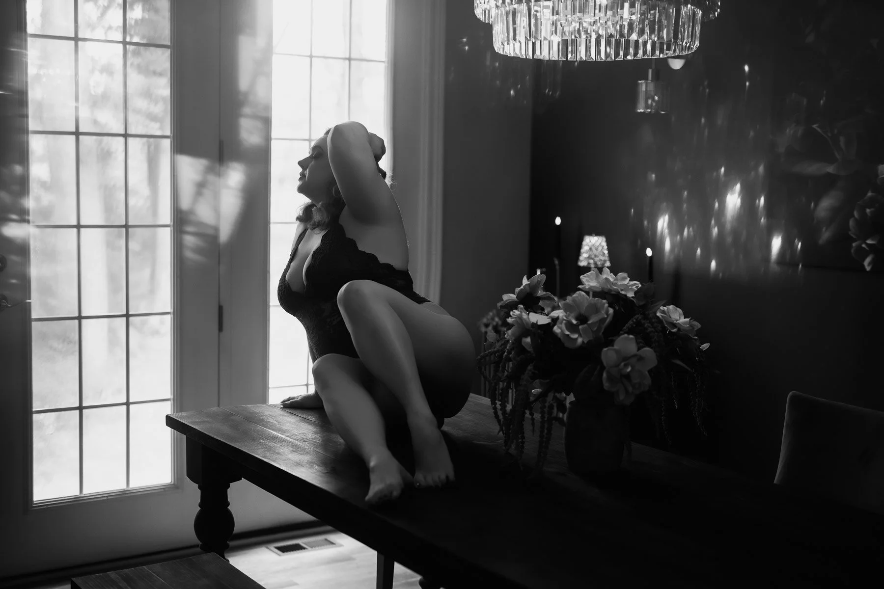 Woman in black lingerie poses on a table in a sunlit room with flowers and a chandelier, captured in black and white.