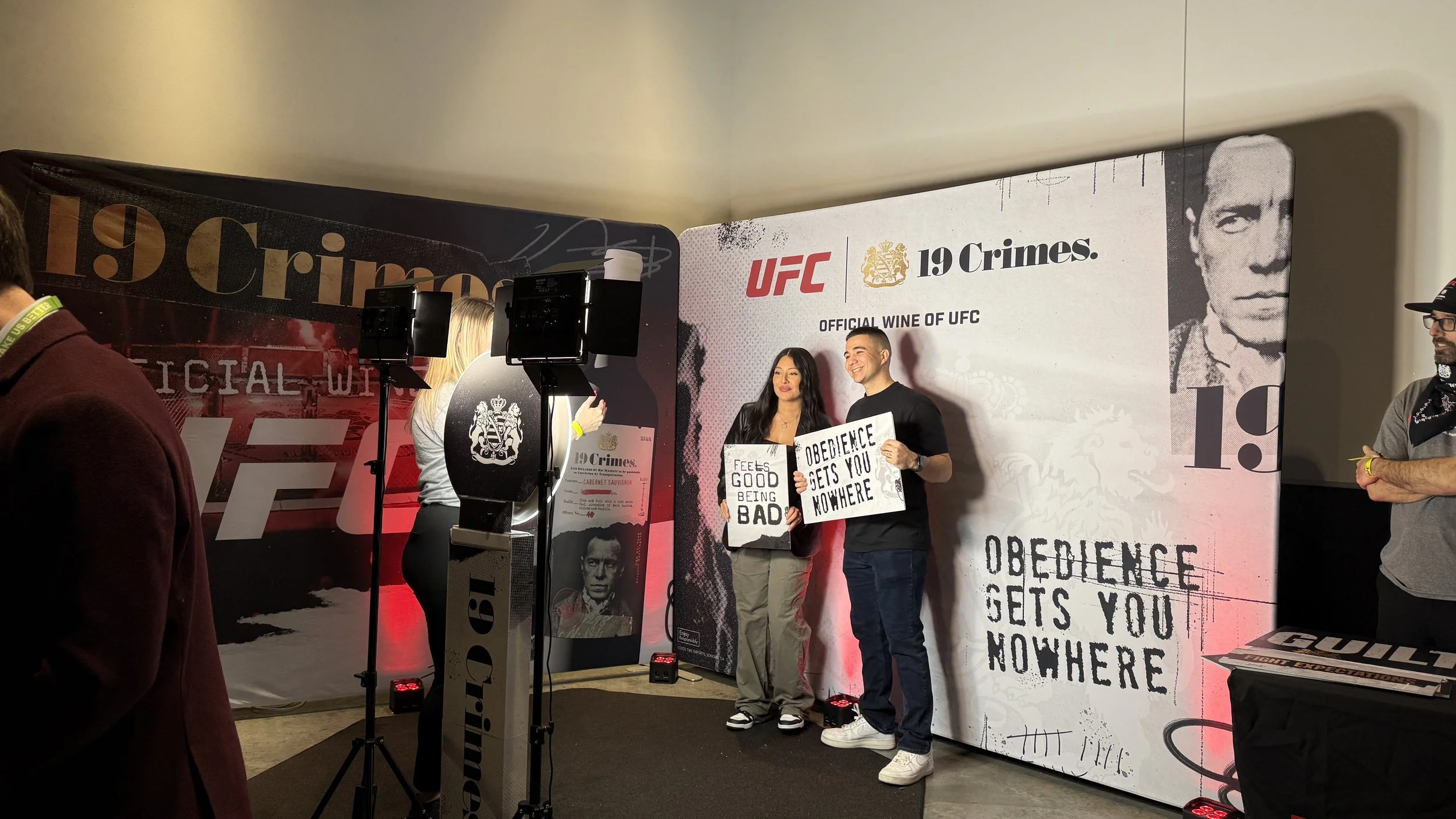 Two people holding signs that say "FEELS GOOD BEING BAD" and "OBEDIENCE GETS YOU NOWHERE" during a UFC event photo shoot with a backdrop featuring the UFC logo and the phrase "19 Crimes" and a stylized black-and-white image of a man.