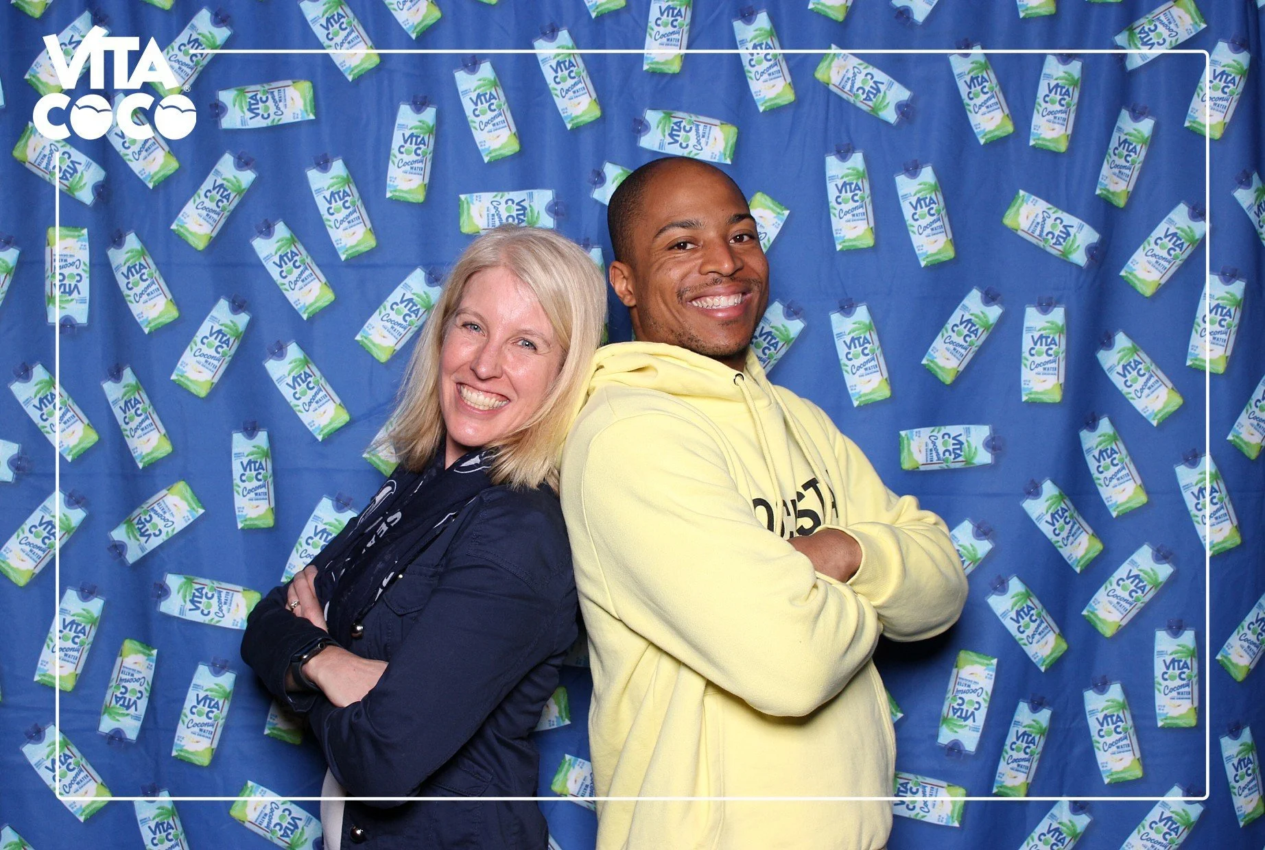 Two people, a woman with blonde hair and a man with short hair, standing back to back in front of a blue backdrop decorated with numerous packages of Vita Coco coconut water. Meet and greet with Tyler Lockett, former Seahawks player.