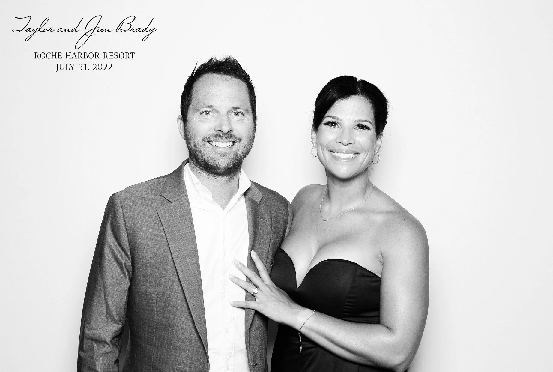 Black and white photo of a smiling man and woman dressed formally, standing close together. The man is wearing a suit and the woman is in a strapless dress, with her hand on the man's chest, showing an engagement ring. Text in the top left corner reads, 'Taylor and Jim Brady, Roche Harbor Resort, July 31, 2022.'