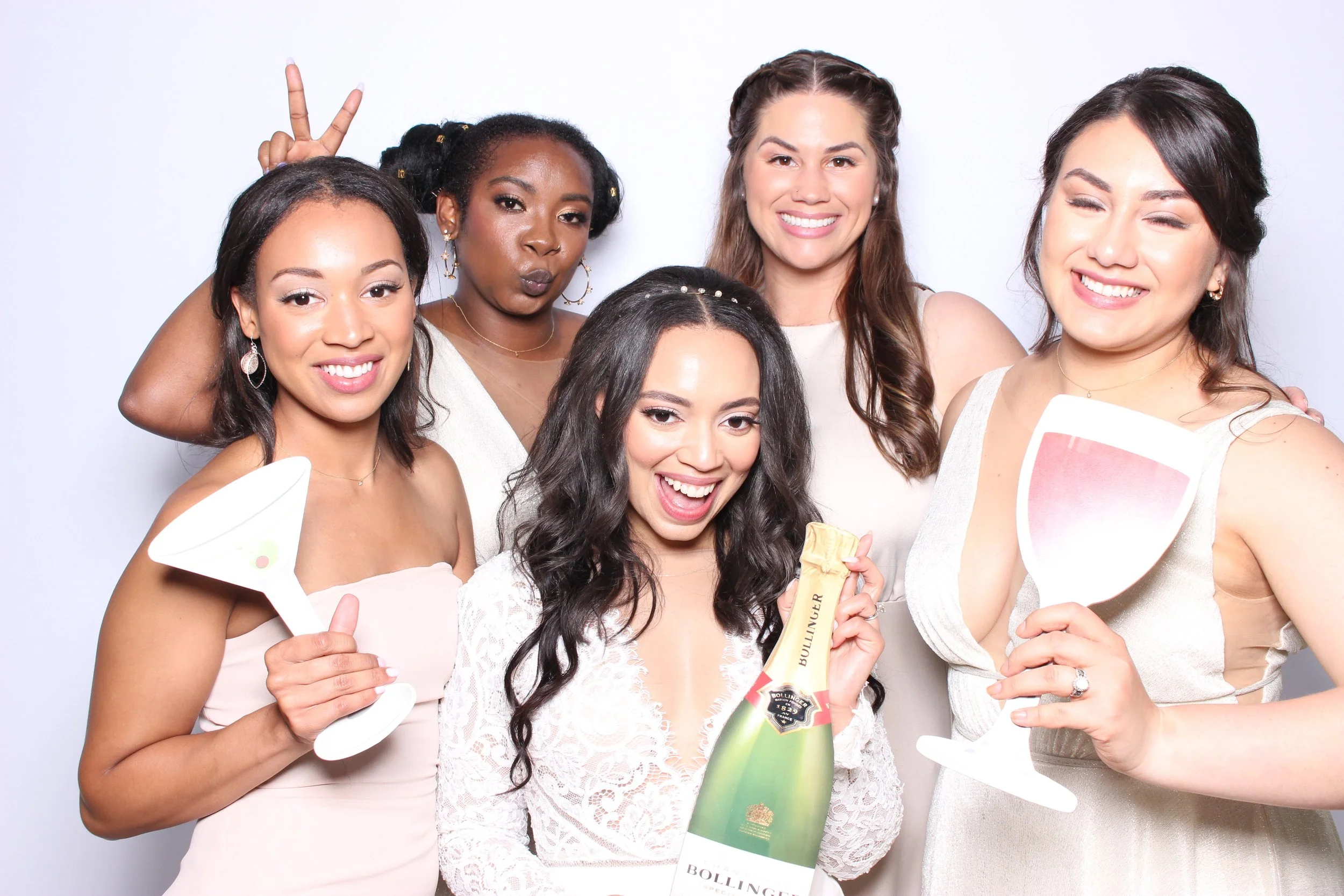 Six women at a celebration, holding champagne glasses and a bottle of champagne, smiling and making playful gestures, dressed in party attire.