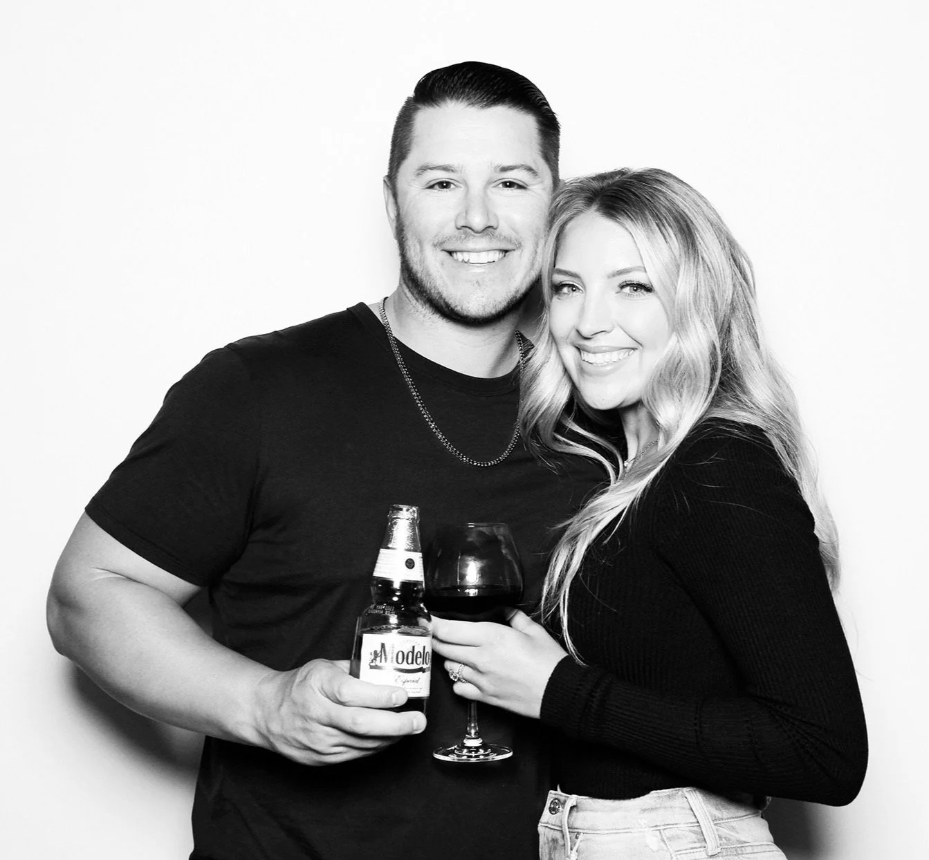 A smiling mariner baseball player and woman stand close together, holding a glass of wine and a bottle of beer, against a plain background.