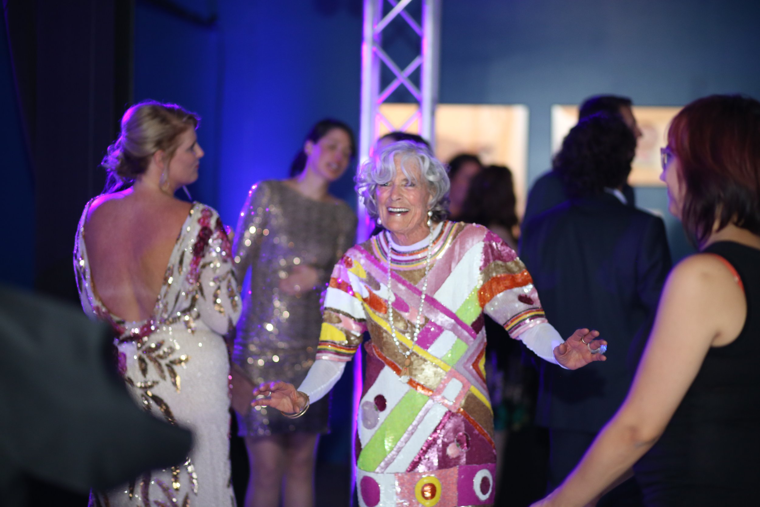 An elderly woman with curly gray hair dancing and smiling at a party, wearing a colorful, geometric-patterned dress with pearl jewelry, while other guests in elegant dresses and suits are in the background.