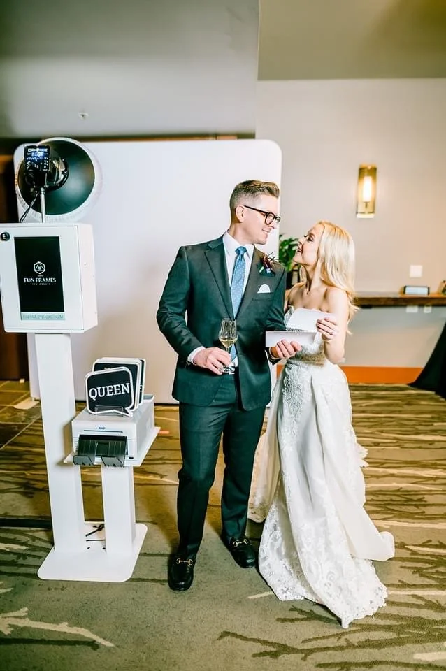 A couple in formal attire at a wedding reception, holding drinks, photo booth prints, and sharing a moment together, with a photo booth station labeled 'QUEEN' in the background.