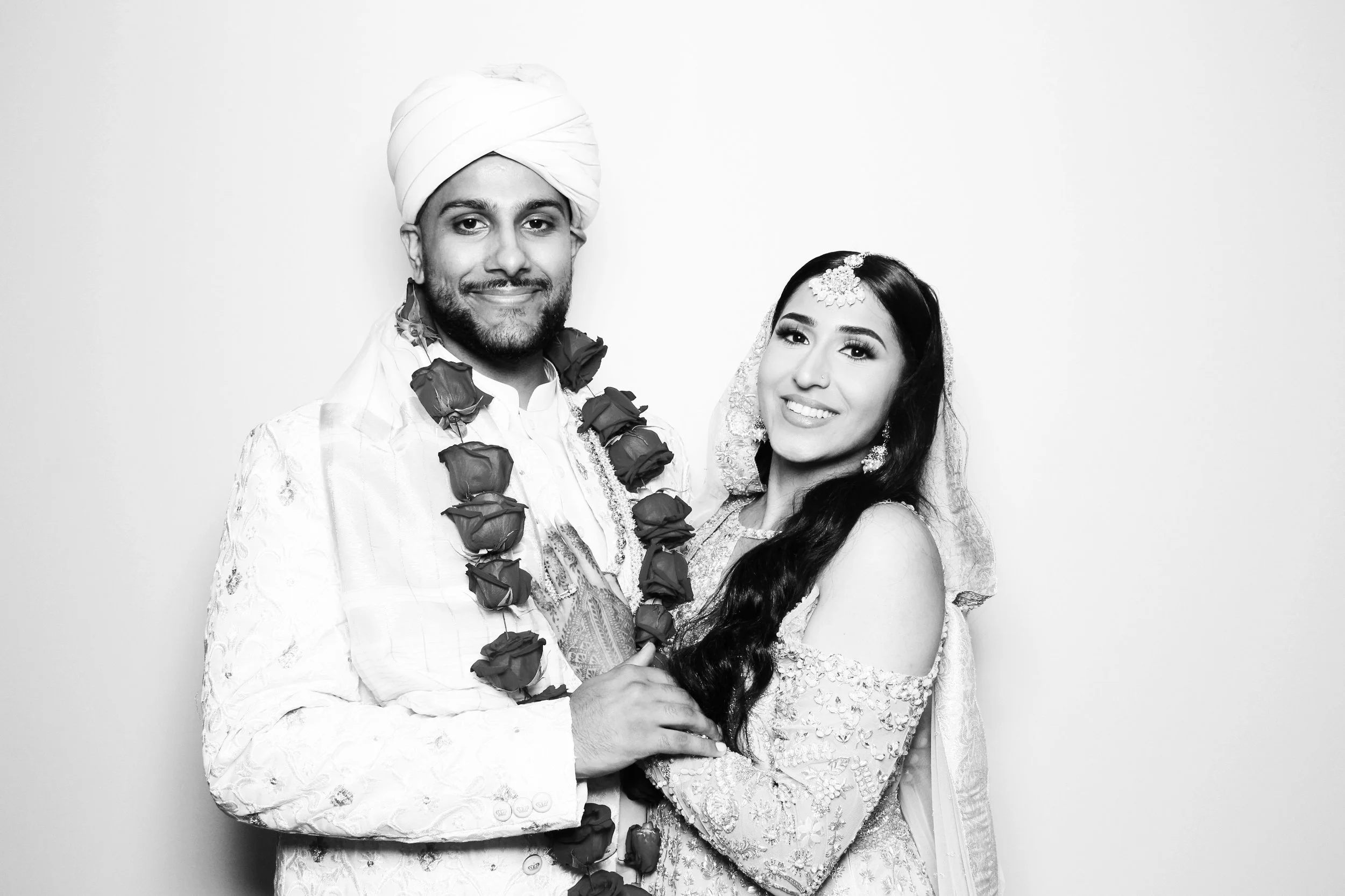 A wedding photo of a couple in traditional Indian attire, with the groom wearing a turban and floral garland, and the bride in an ornate dress and jewelry, smiling and posing together.