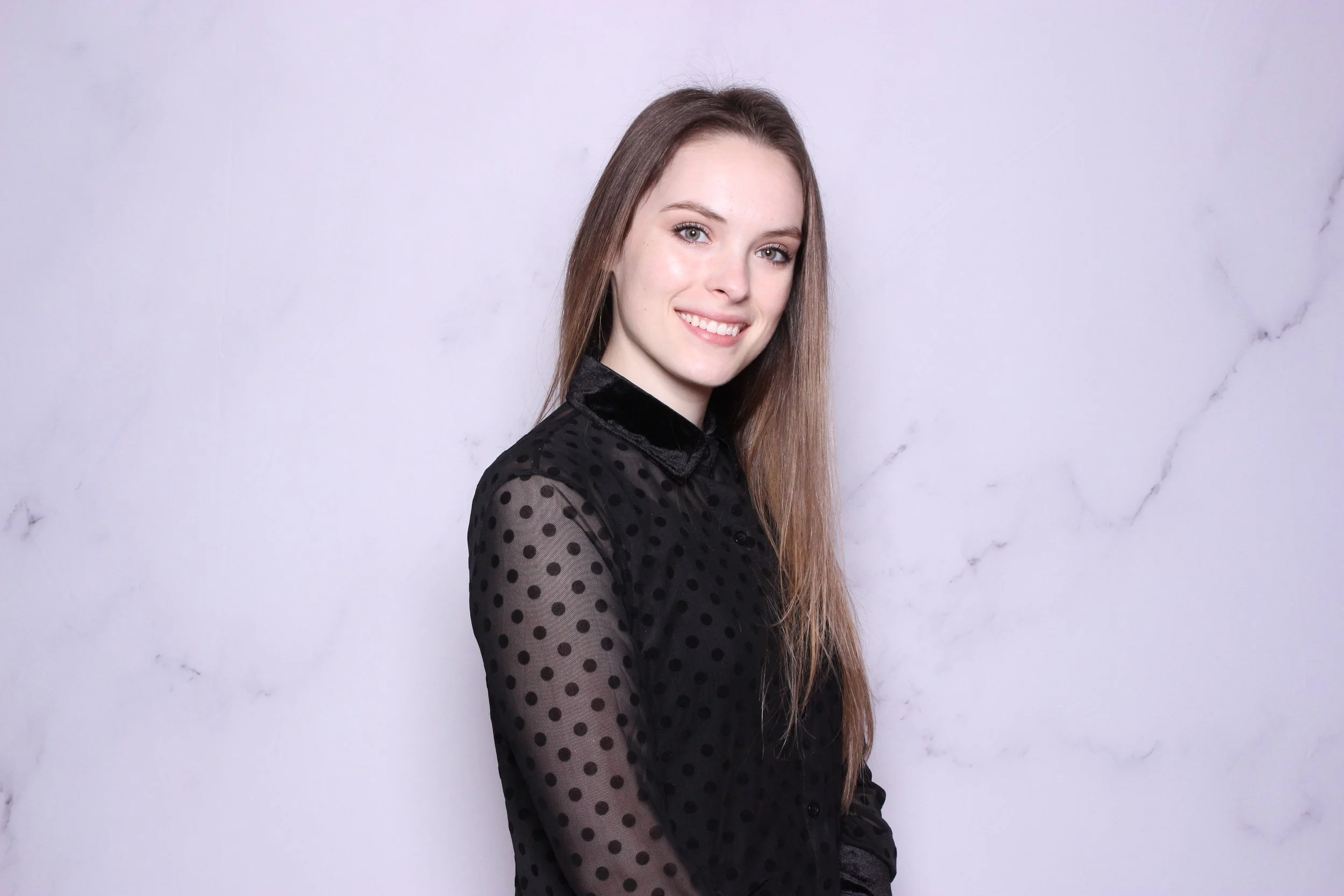 A young woman with long brown hair, smiling, wearing a black polka dot sheer blouse, standing against a light gray marble background.