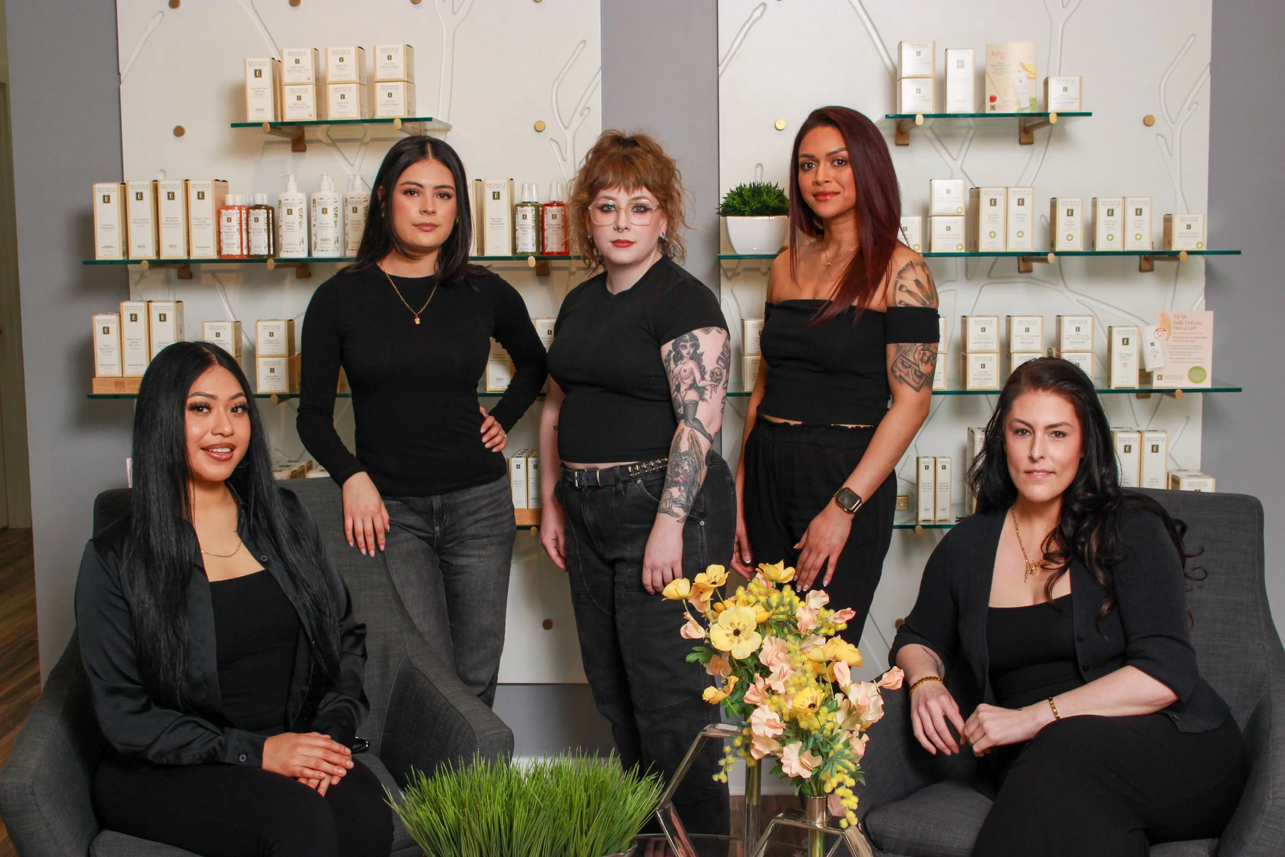 Five women in black clothing posing in a room with shelves of skincare products behind them, two sitting on gray couches, three standing in the middle, with flowers and greenery on a table in front at their beauty salon.