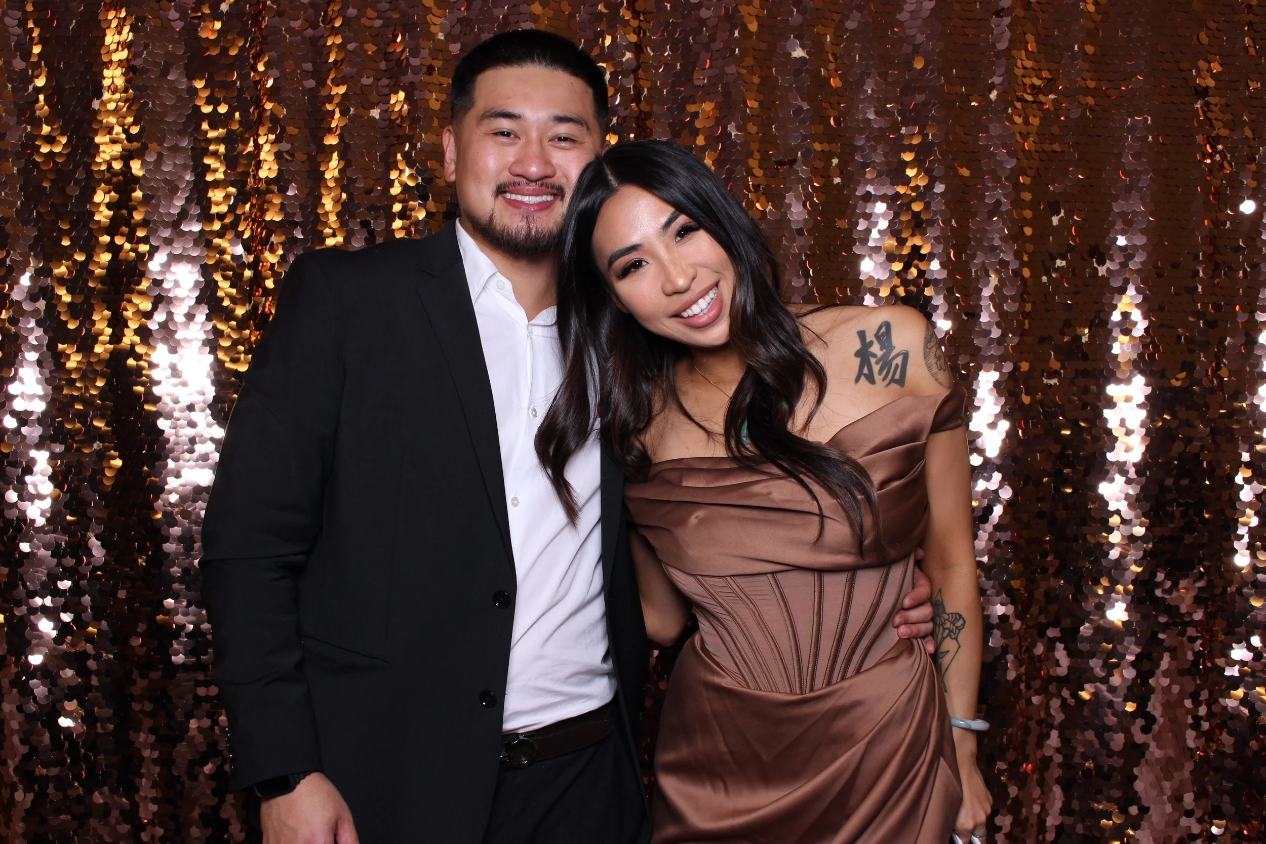 A smiling man in a black suit and white shirt standing next to a smiling woman in a brown dress with a tattoo on her shoulder, against a rose gold sequin backdrop.
