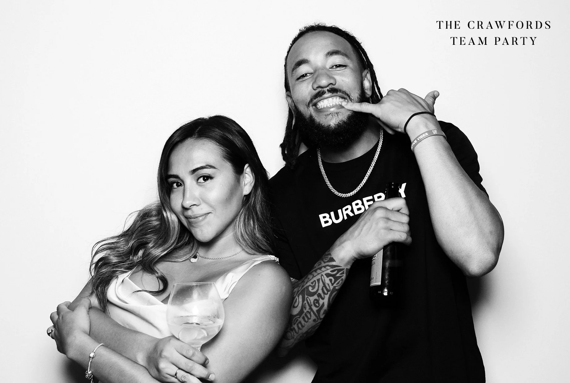Black and white photo of a Kathy and Mariner Baseball Player J.P. Crawford at a team party, with the woman holding a glass of wine and the man holding a beverage bottle, both smiling and posing playfully.