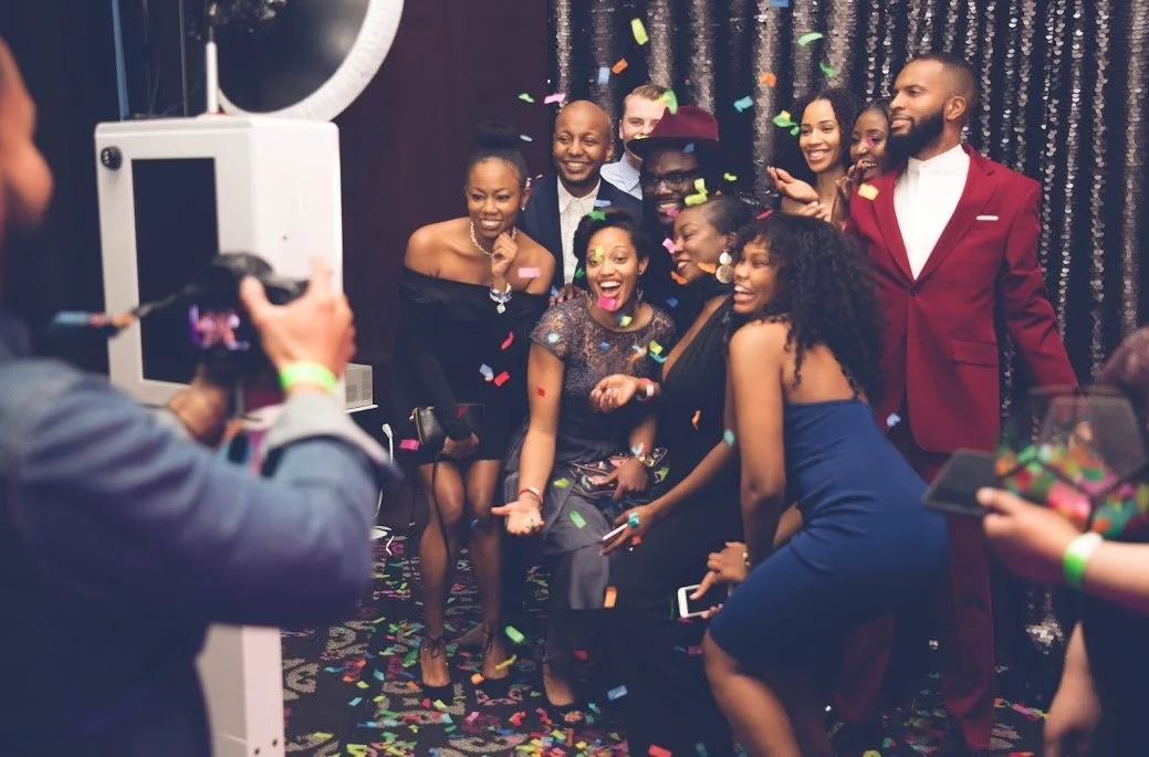 A group of people dressed in formal attire celebrating with confetti in front of a photo booth.