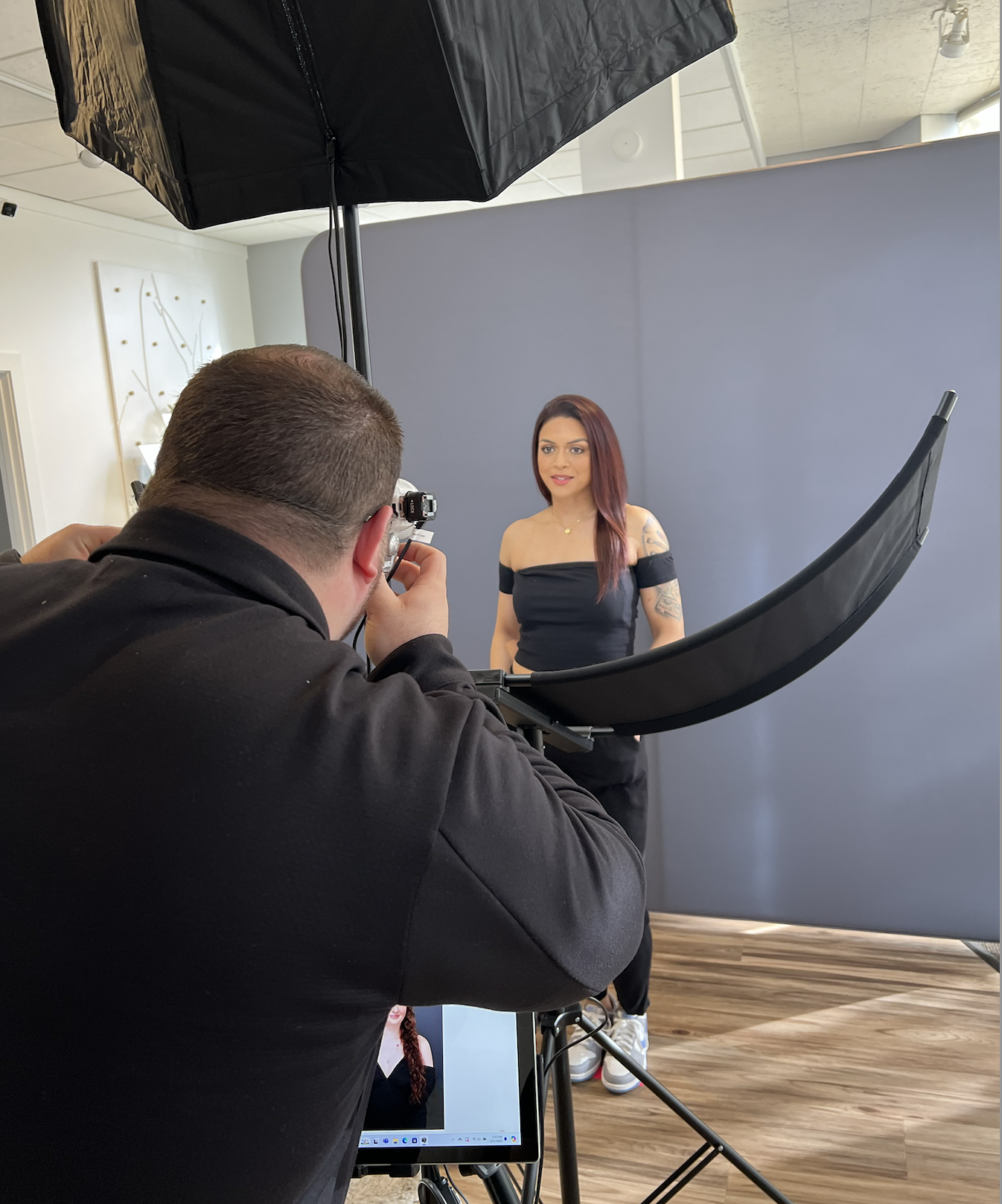 Photographer taking a picture of a woman with tattoos standing in front of a gray backdrop in a studio.