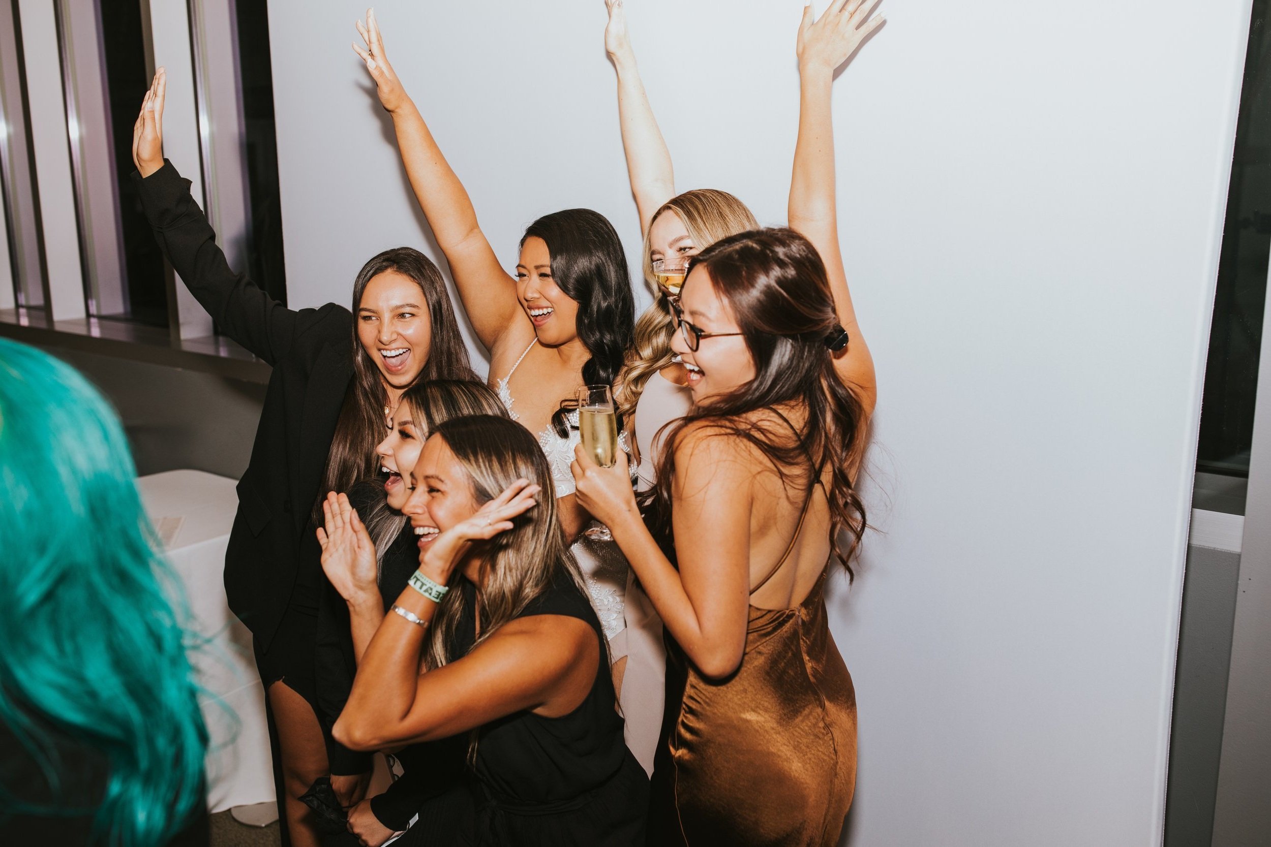 Group of six women celebrating in the photo booth, smiling and laughing at a party, one holding a glass of champagne, with some raising their arms in joy. Wedding at Chihuly Garden and Glass, Seattle.