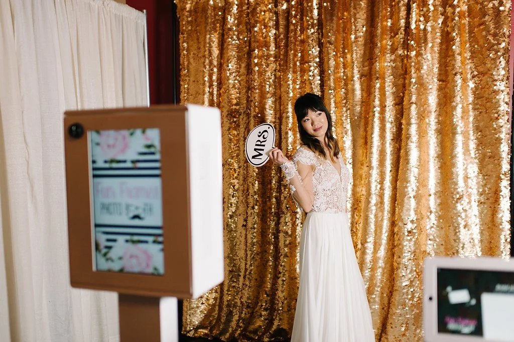 A woman in a wedding dress holding a 'MRS' sign, standing in front of a gold sequin backdrop at a photo booth.