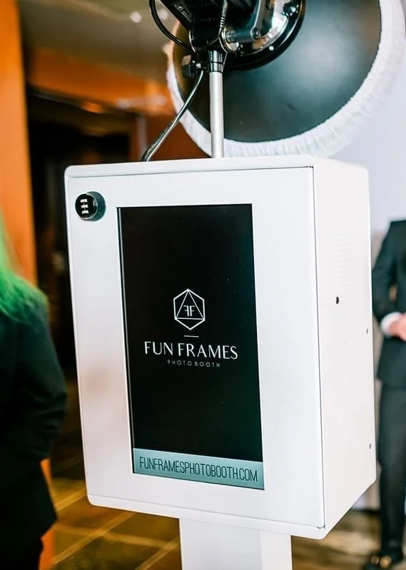 A photo booth company named "Fun Frames" with a white exterior and a digital screen displaying the logo and website, located indoors at an event.