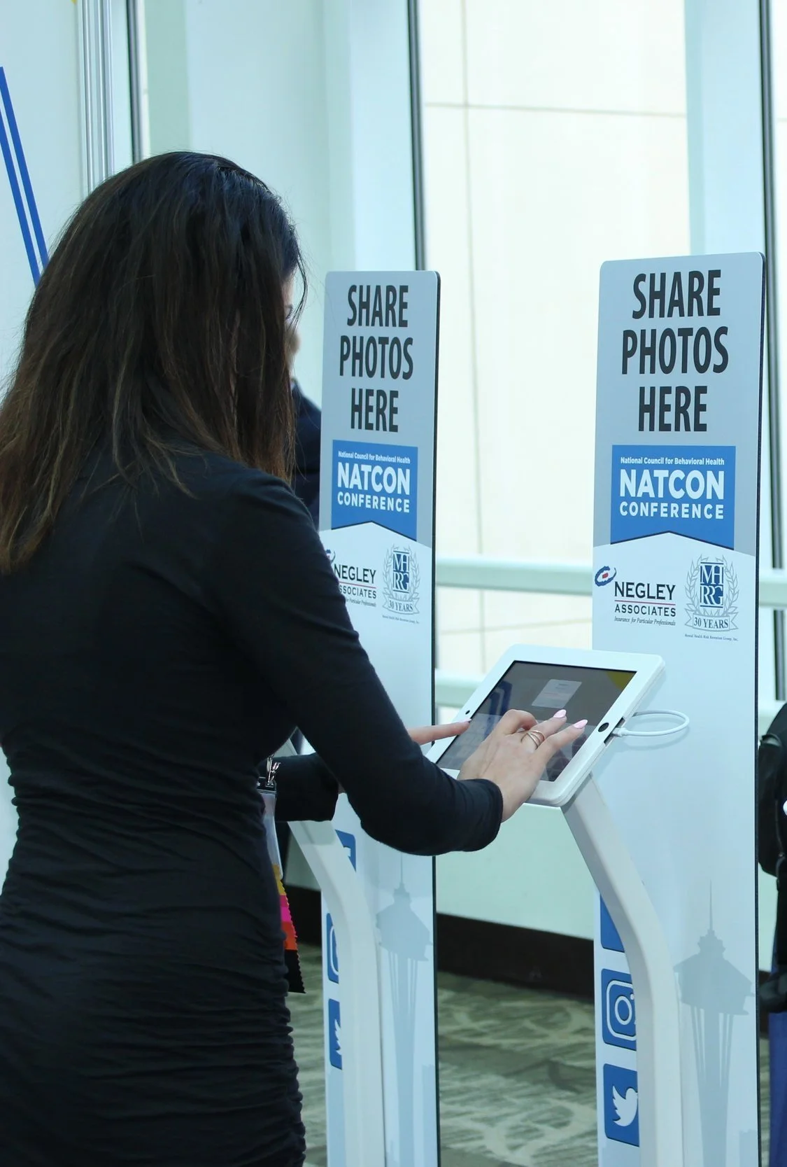 Woman using a digital kiosk at a conference with signs that say 'Share Photos Here' for the NATCON Conference.