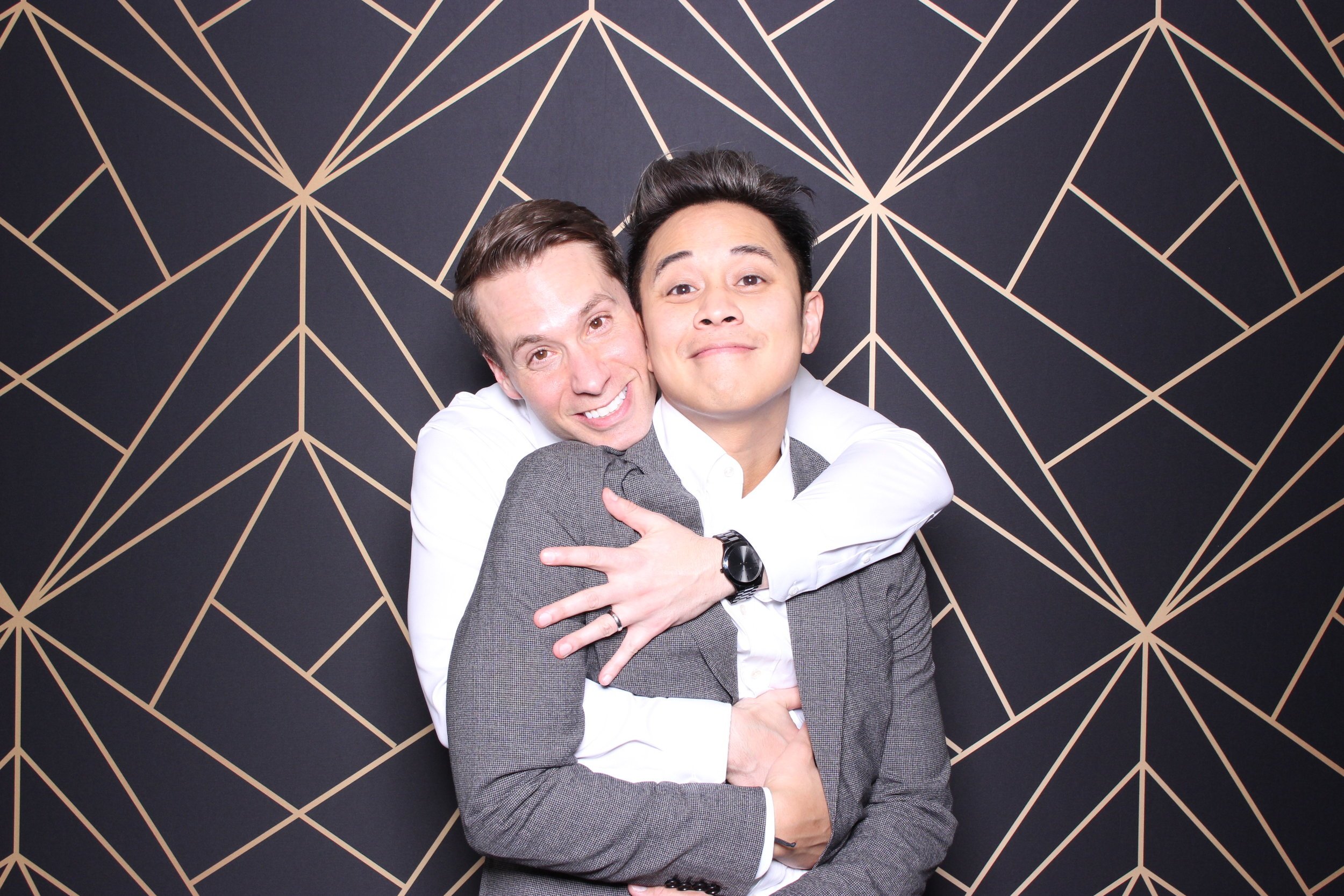 Two men embracing, one with his arms around the other, standing against a geometric patterned black and gold background.