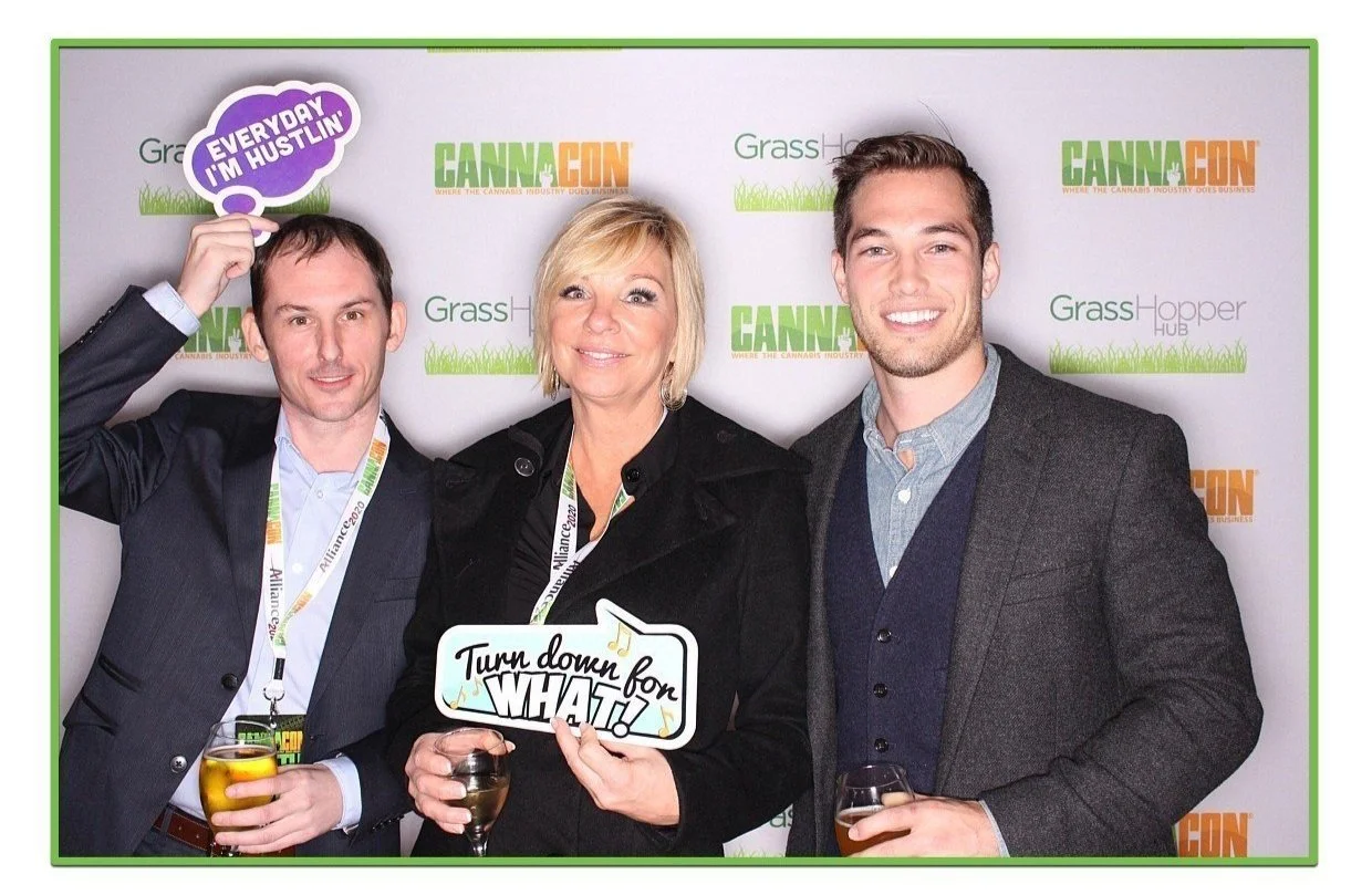 Three people at a cannabis industry event standing in front of a backdrop with the words "Grass Hopper Hub" and "CannaCon." The person on the left is holding a sign that says "EVERYDAY I'M HUSTLIN." The woman in the middle is holding a drink and a sign that says "Turn down for WHAT." The man on the right is smiling, holding a drink, and wearing a dark jacket over a light shirt and sweater.