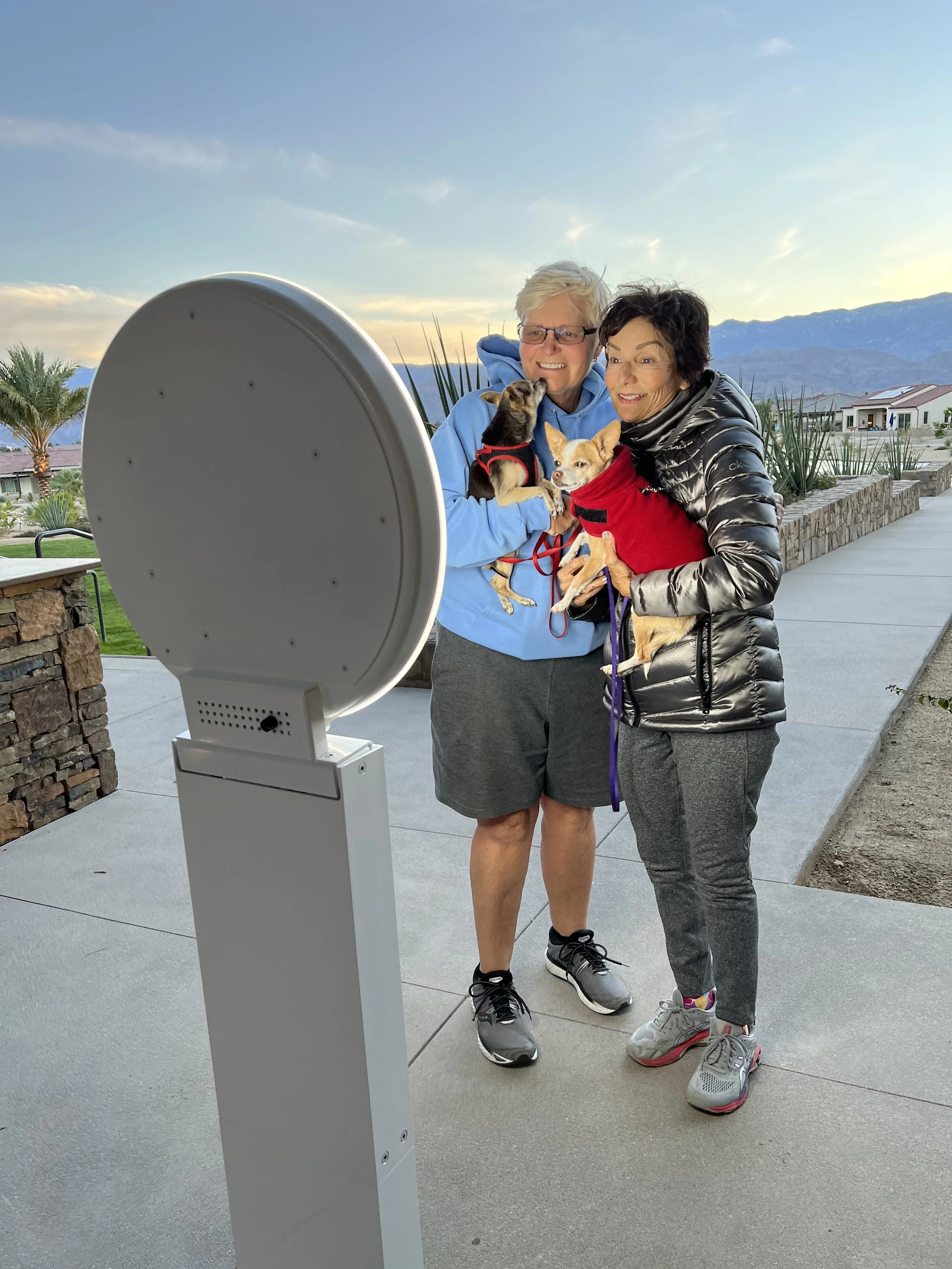 Two women holding small dogs in a selfie photo booth and standing outdoors in Rancho Mirage, CA with mountains and a cloudy sky in the background.