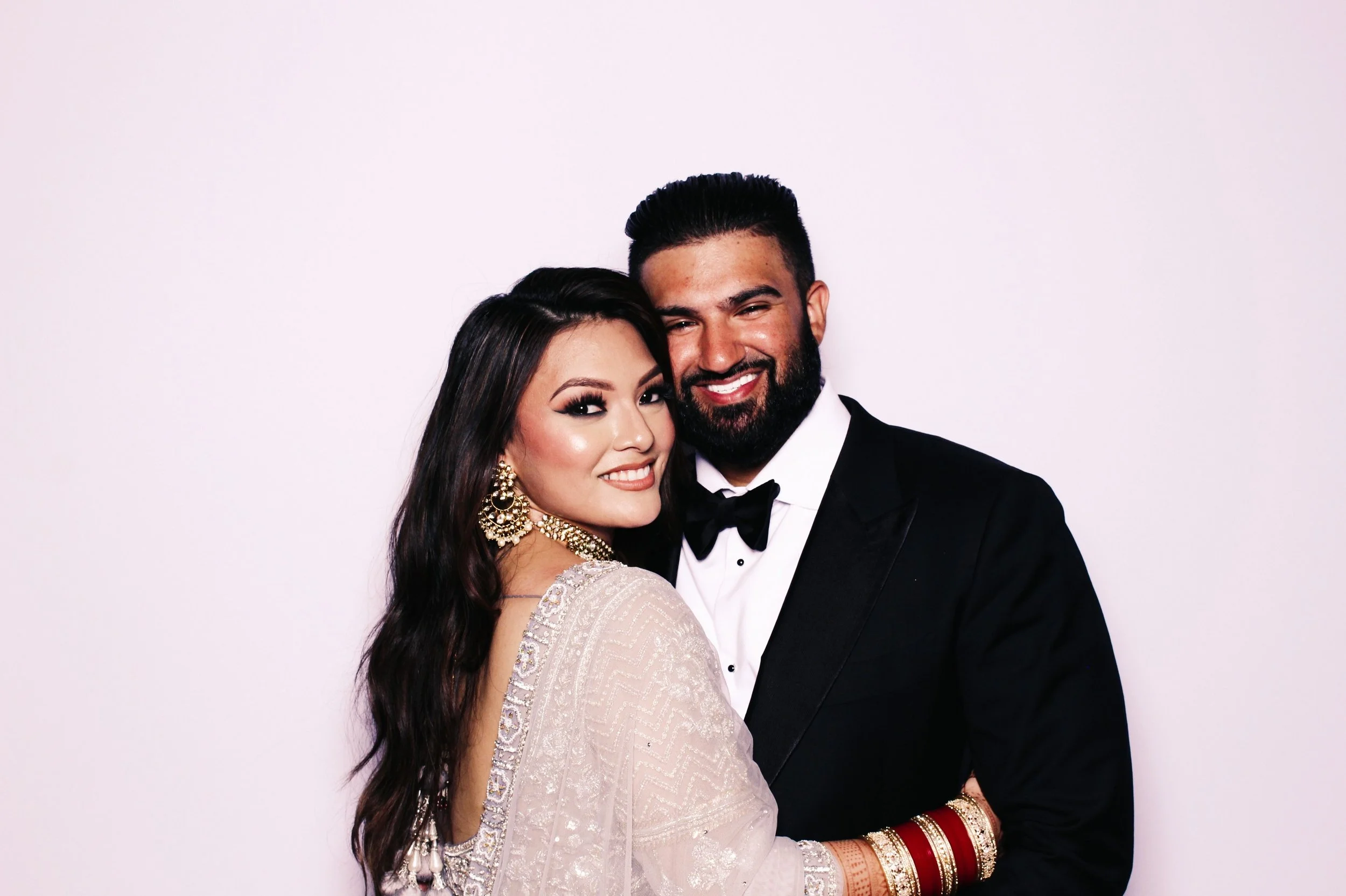 A smiling woman and man dressed in formal attire, the woman wearing traditional Indian clothing and jewelry, and the man in a black tuxedo with a bow tie, posing together against a plain white background.