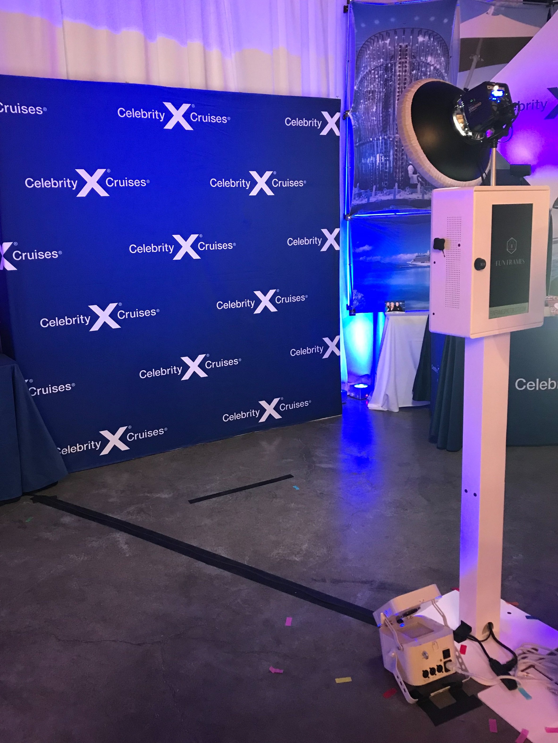 Photo of a photo booth setup with a blue backdrop that says "Celebrity Cruises" and "Celebrity x Cruises" in a repeating pattern. The photo booth includes a camera on a tripod, various lighting equipment, and a stand with a screen.