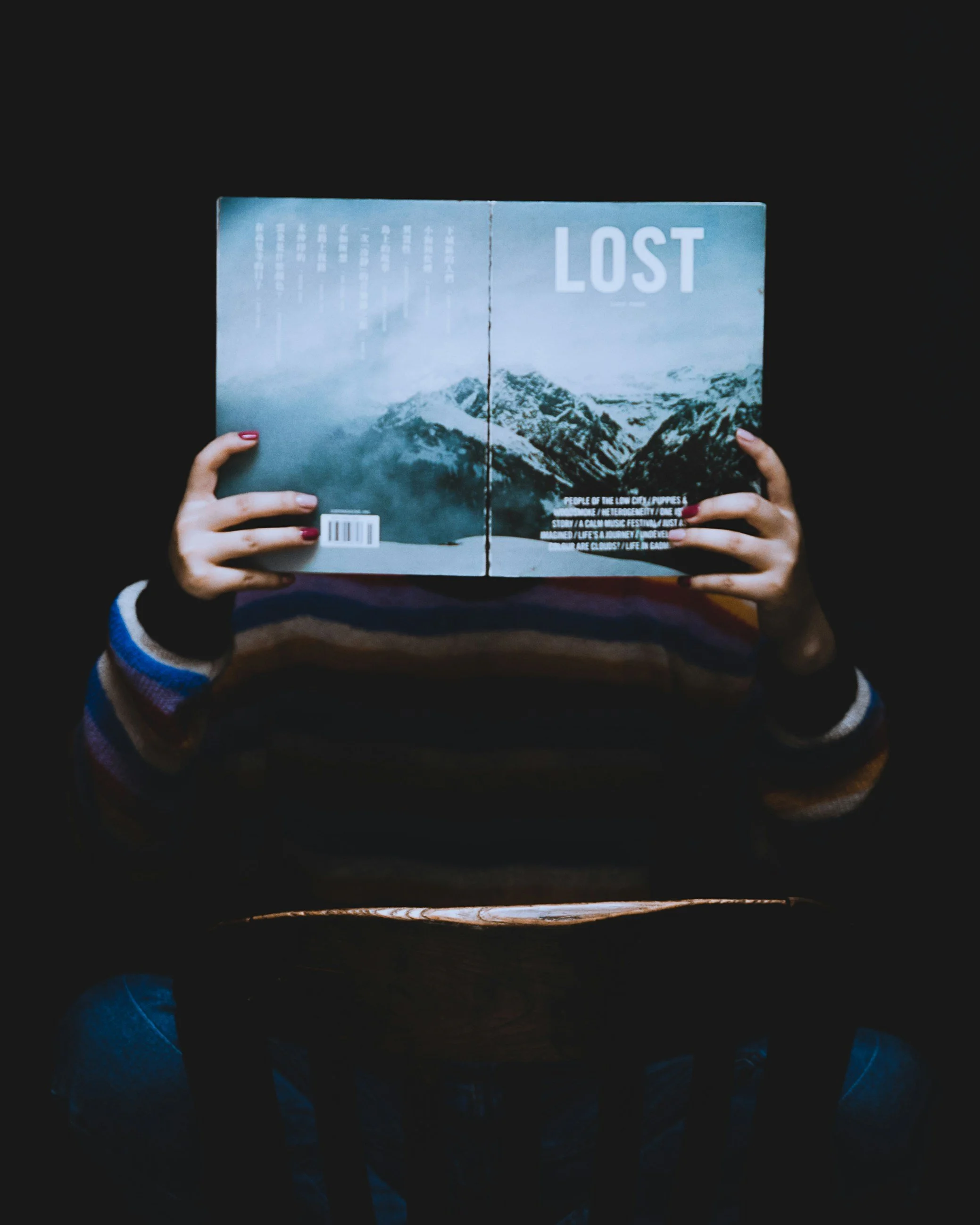 Person hält ein Buch mit dem Titel 'LOST' vor das Gesicht, sitzt an einem dunklen Tisch, trägt einen bunten Pullover, umgeben von Dunkelheit.