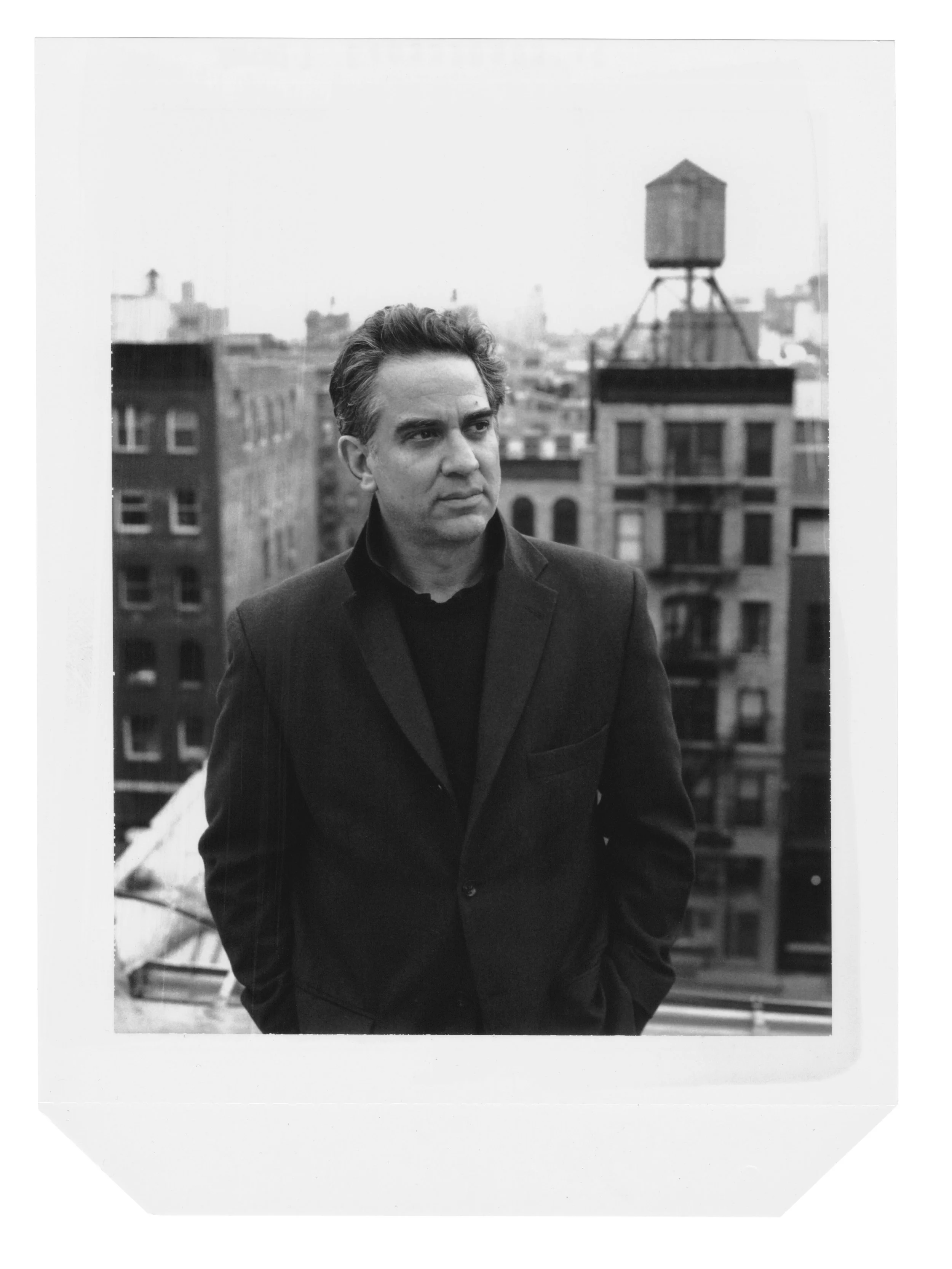 A Polaroid photograph of artist Tom Slaughter is shown. Slaughter wears a suit jacket while he stands on a rooftop in the New York City and looks out to the right of the camera.