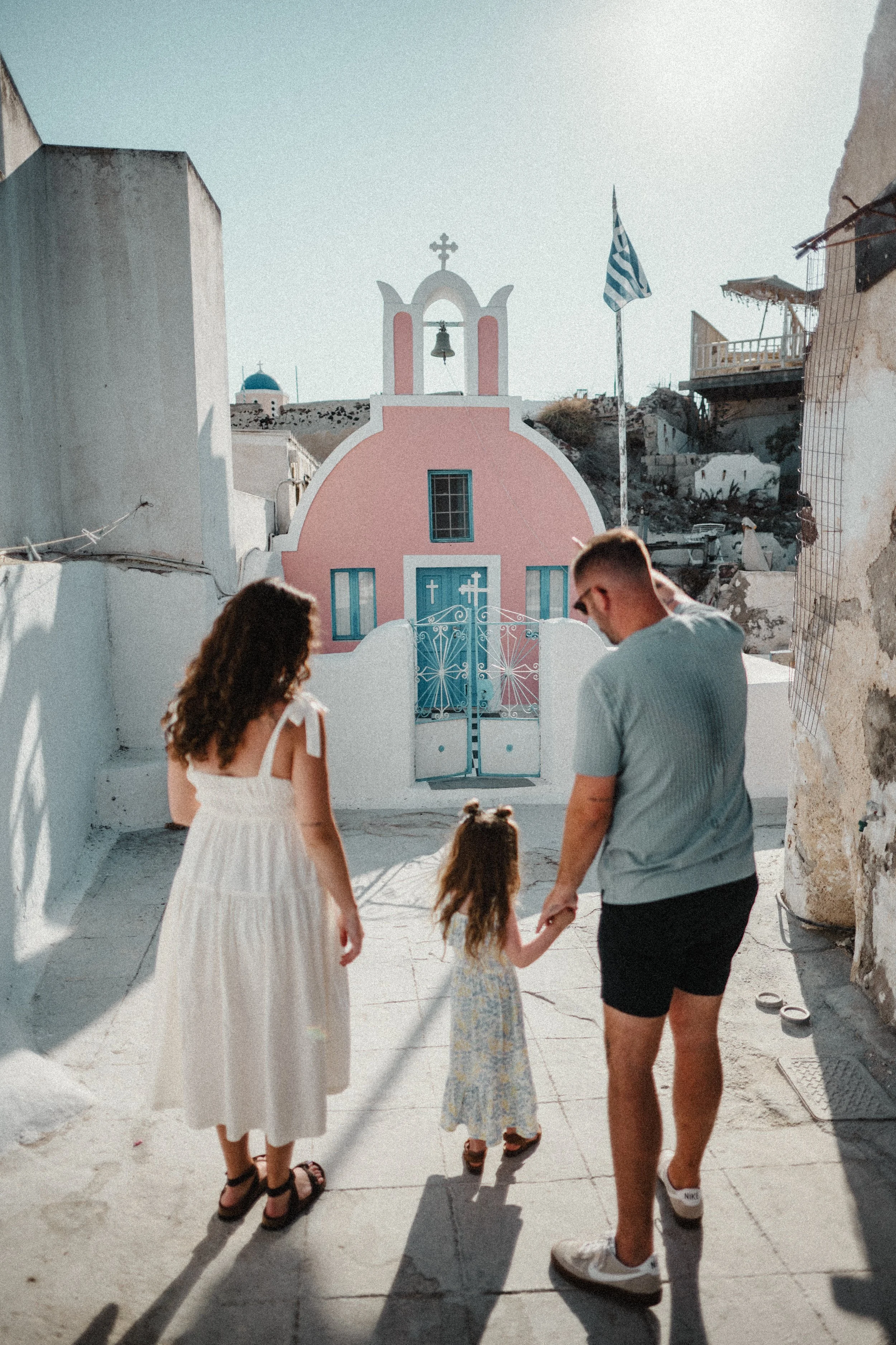 A young family holding hands while strolling toward a charming pink Greek chapel with teal ironwork gates and a bell tower, the Greek flag flying above — candid and lifestyle Santorini family photography session through the village alleyways.