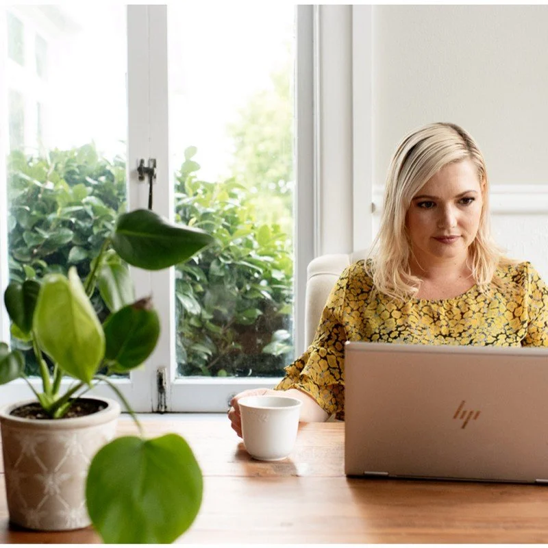 A woman with blonde hair working on a laptop at a wooden table near a window, with a potted plant and a cup on the table.