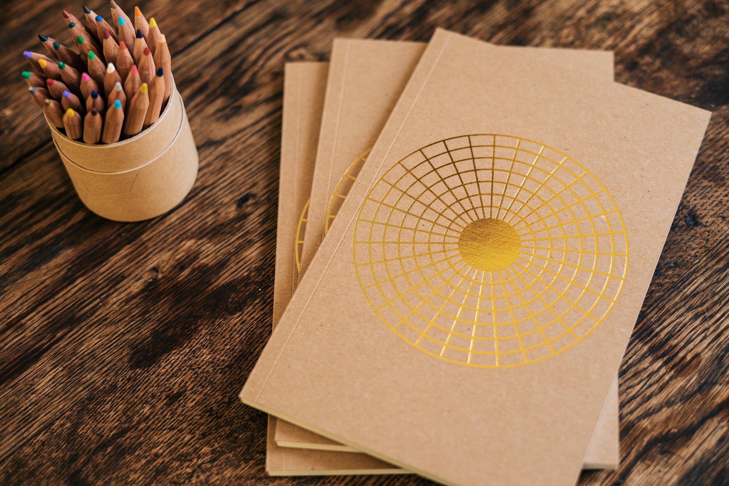 3 journals on table with colored pencils in a brown cardboard tube. 