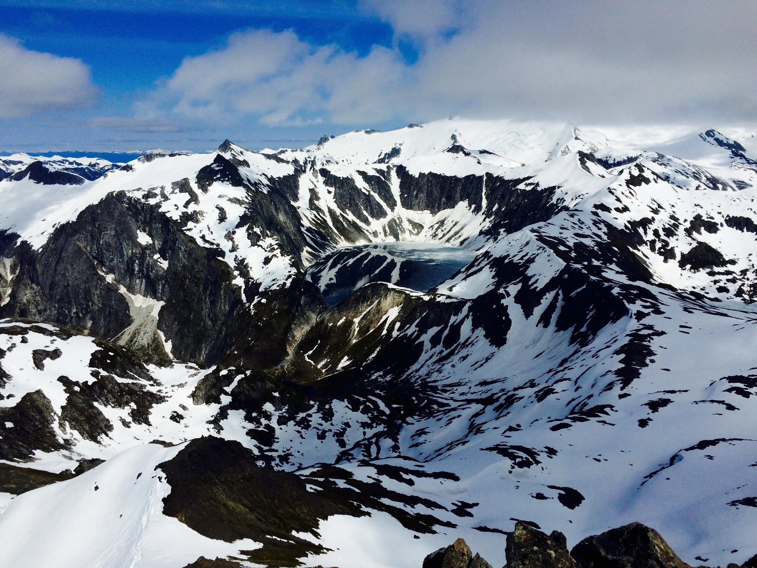 Snow-covered mountain peaks with rocky ridges and a small frozen lake in the valley beneath, under a partly cloudy sky.