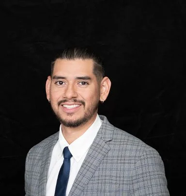 A man with dark hair and a beard, wearing a gray checkered suit, white shirt, and blue tie, smiling in front of a black background.