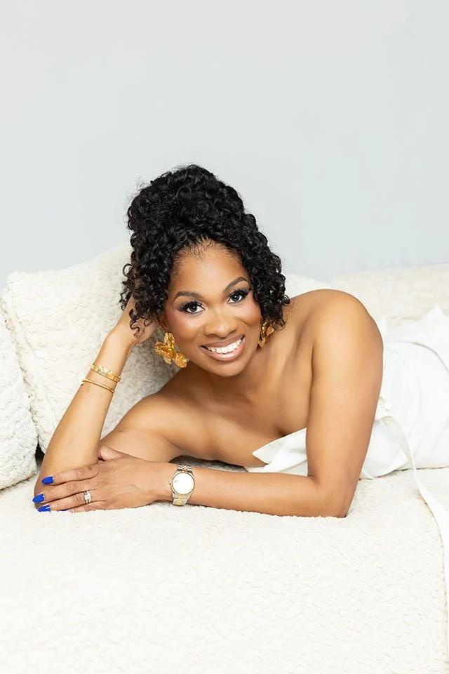 A smiling woman with curly black hair, gold earrings, a white strapless dress, blue nail polish, a gold watch, and bracelets, lying on a white bed with a white pillow.