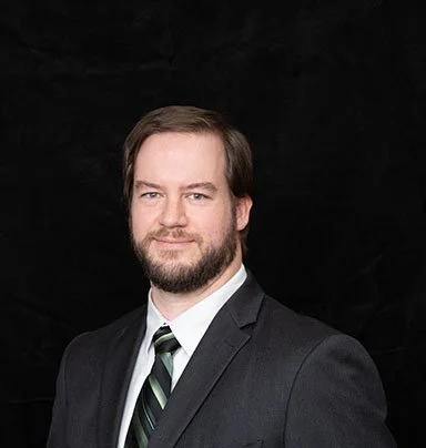 Professional headshot of a man in a dark suit with a striped tie, posing against a black background.
