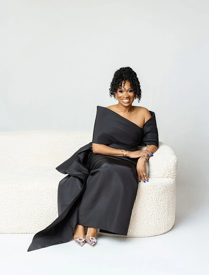 A woman dressed in a black off-shoulder gown sitting on a white textured sofa against a plain white background, smiling at the camera.