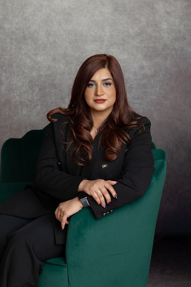 A woman with long, wavy auburn hair, wearing a black blazer, sitting on a green armchair against a gray textured background.
