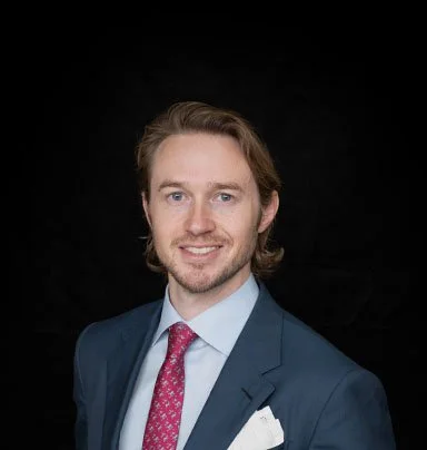 A professional man with shoulder-length brown hair, wearing a navy suit, pink tie, and white shirt, smiling against a black background.